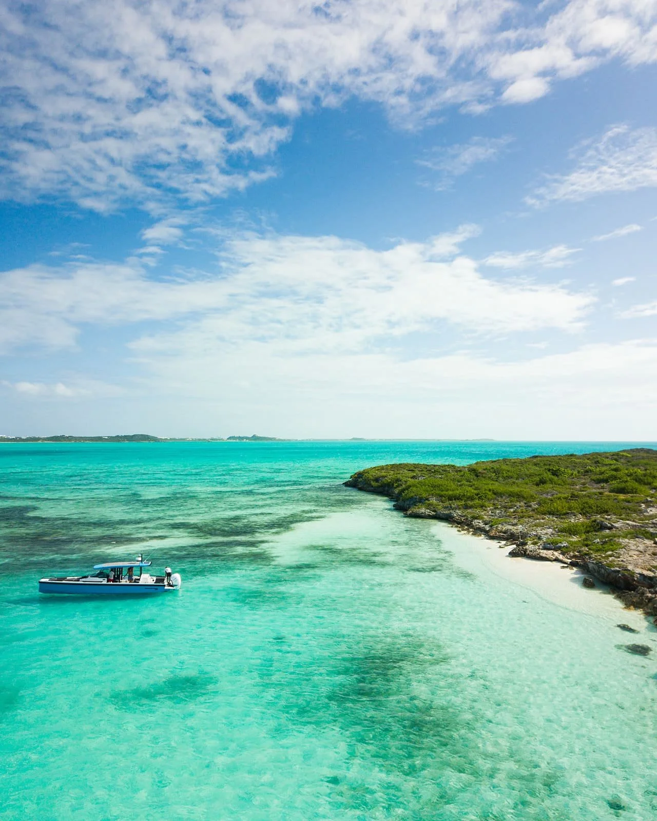 A boat floating on clear turquoise water near a rocky shoreline with green vegetation on a sunny day with partly cloudy sky.