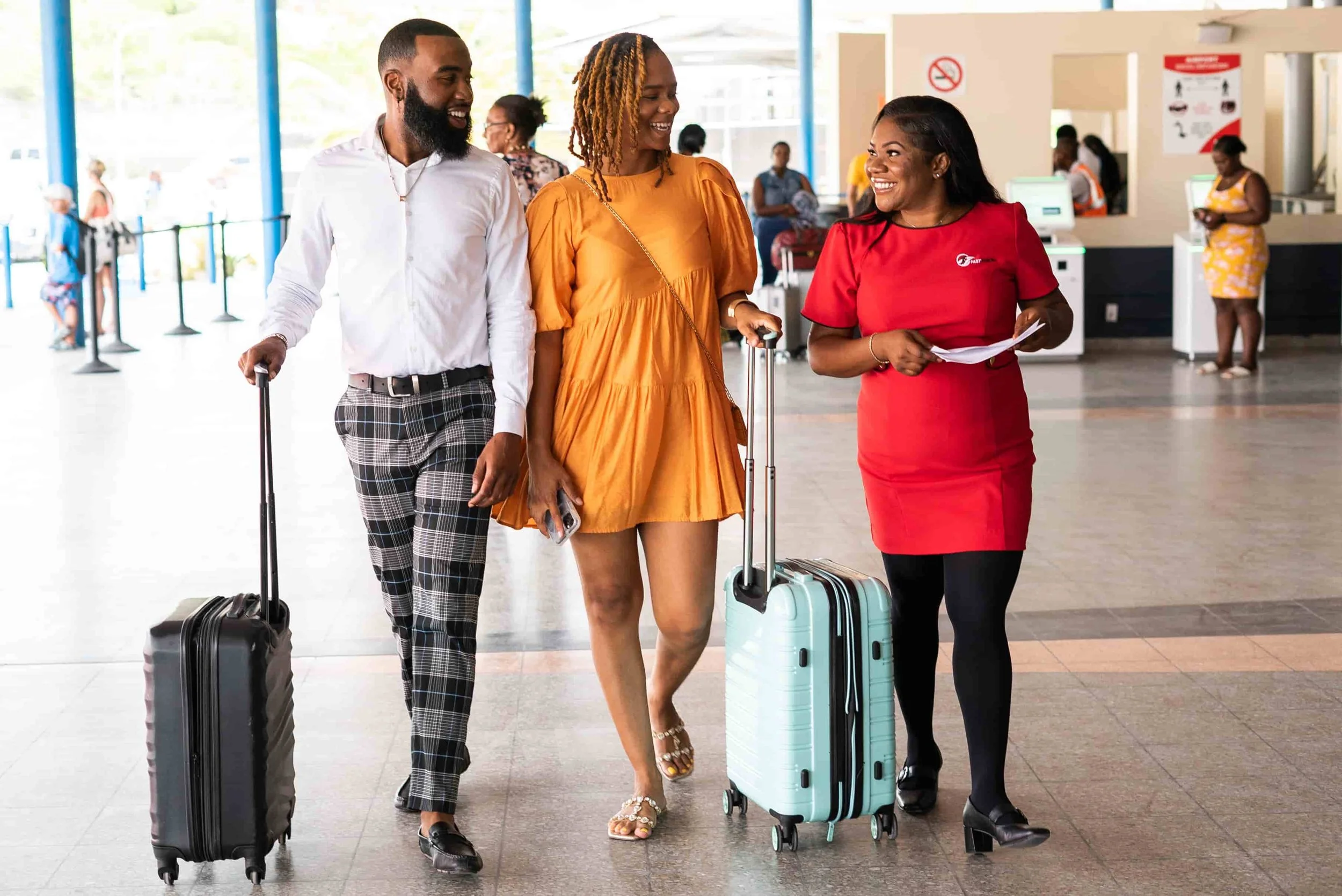 Three people walking through an airport, talking and smiling. Two are pulling rolling suitcases, and one is holding papers and standing next to a suitcase. The background shows other travelers, counters, and large windows.