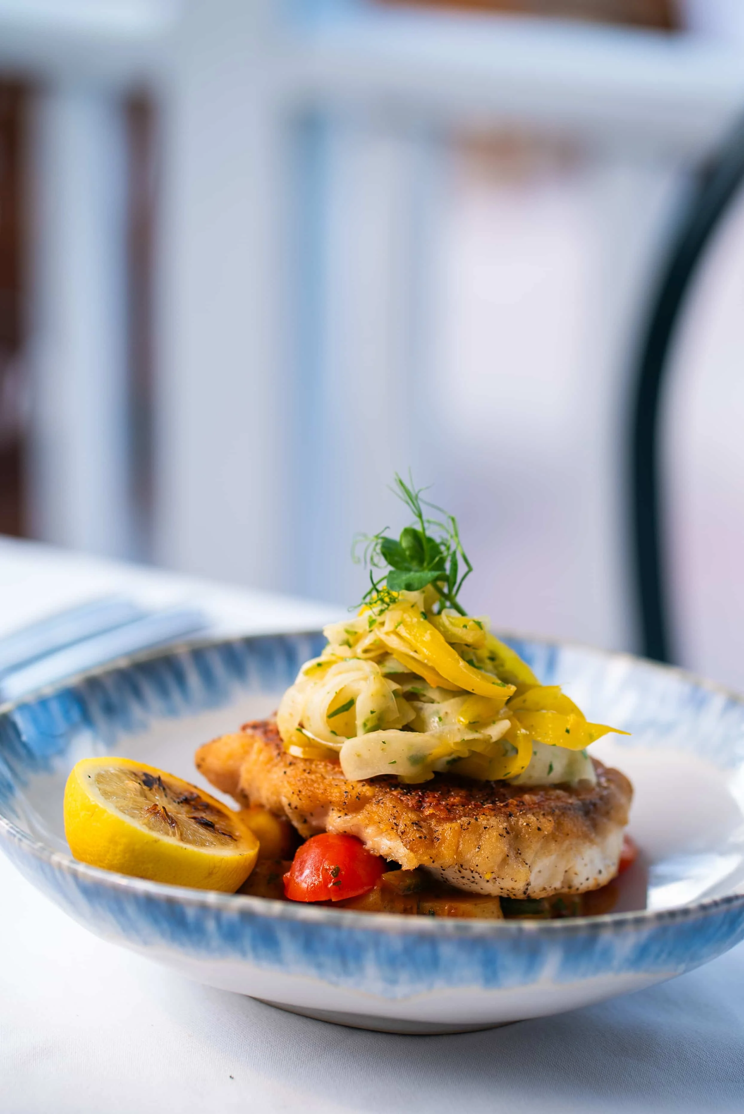 A plated dish featuring a piece of seasoned fried fish topped with fennel and yellow pepper slaw, garnished with microgreens, served with a lemon wedge on a blue-rimmed plate.