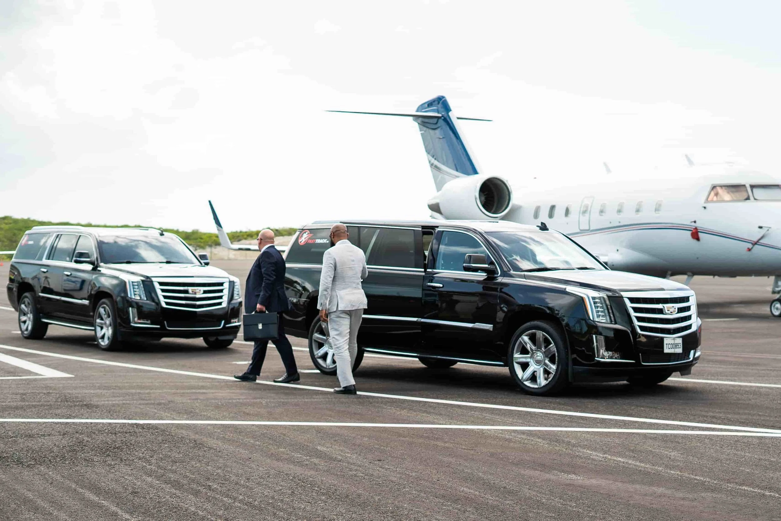 Two black Cadillac limousines parked on an airport tarmac with two men in suits walking toward the limousines. An private jet is visible in the background.