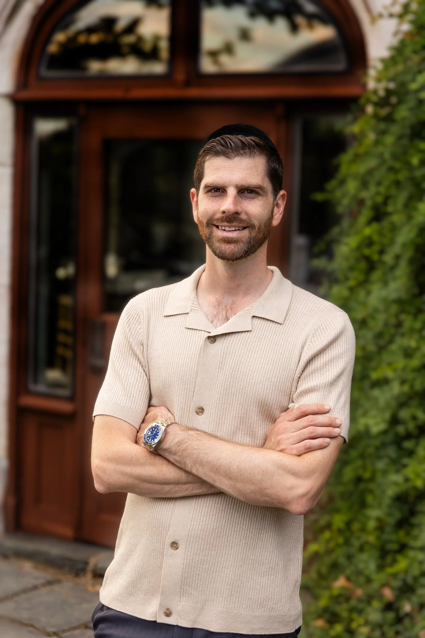 A man with brown hair and a beard stands outside with arms crossed, smiling, wearing a beige short-sleeved polo shirt and a wristwatch, in front of a house with a wooden door and green shrubbery.