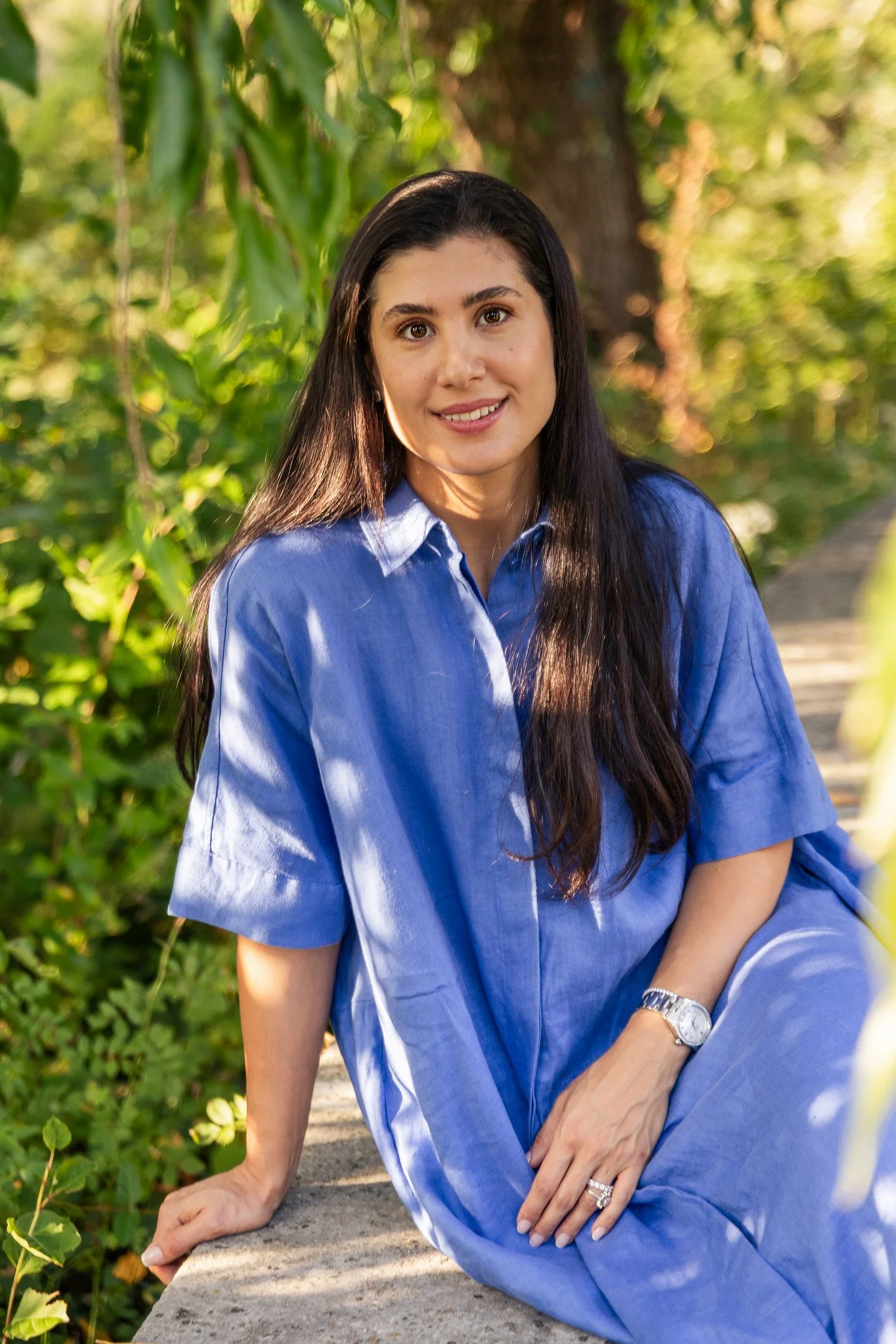 A woman in a blue dress sitting outdoors on a concrete ledge, smiling with trees and greenery in the background.