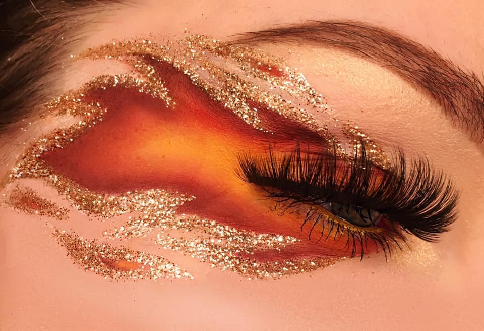 Close-up of a closed eye with orange and gold glitter eyeshadow, black long eyelashes, and well-groomed eyebrows.