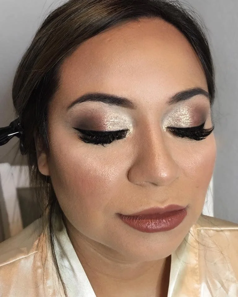Close-up of a woman with styled hair and glamorous makeup, including shimmery silver eyeshadow, thick black eyeliner, long false eyelashes, and lipstick in a warm brown shade.