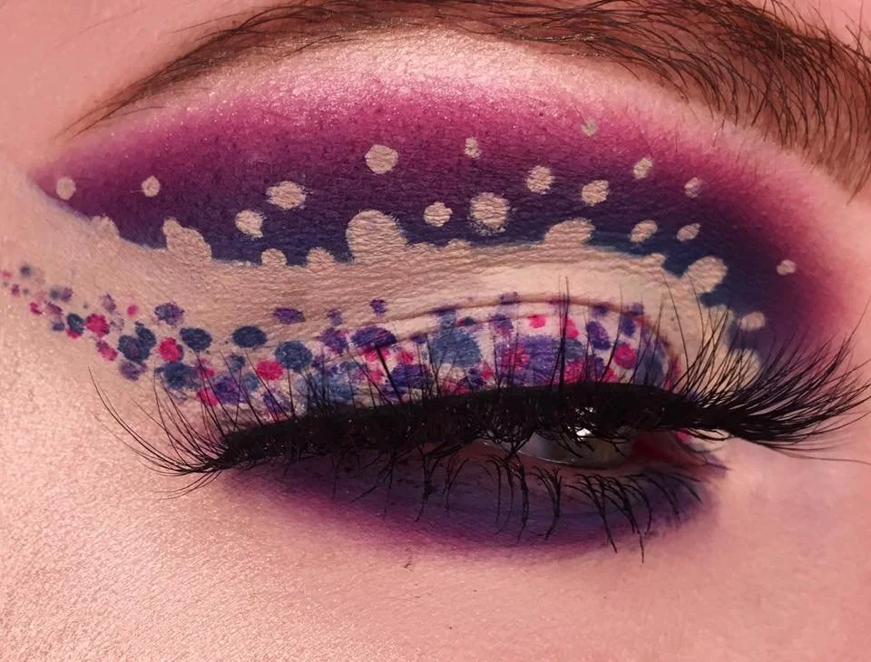 Close-up of eye with vibrant, colorful eye makeup featuring pink, purple, and blue shades, with white paint dots on eyelid and around eye, and long black eyelashes.