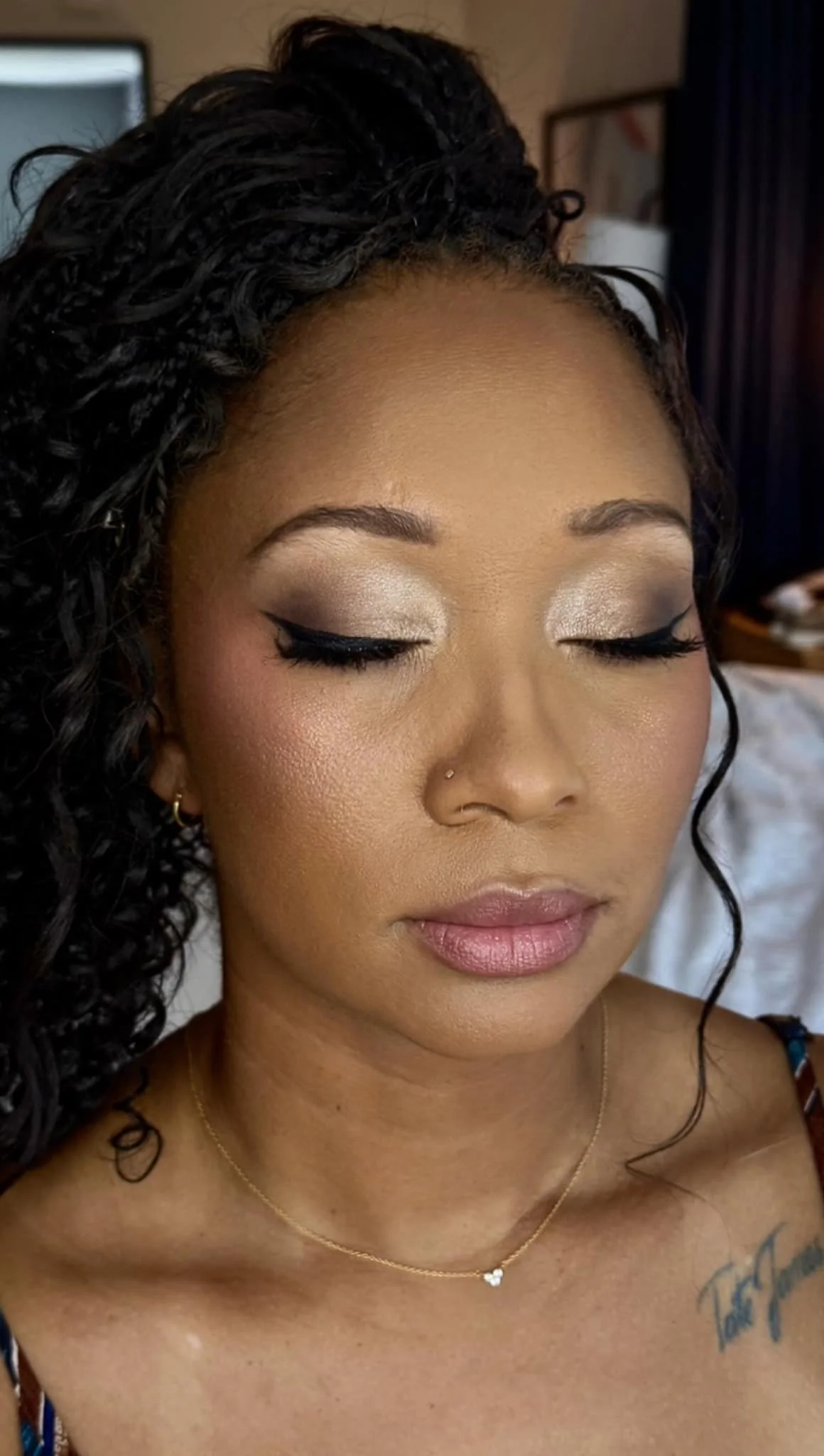 Close-up of a woman with curly black hair and makeup, wearing a gold necklace, small earrings, and a tattoo on her shoulder.