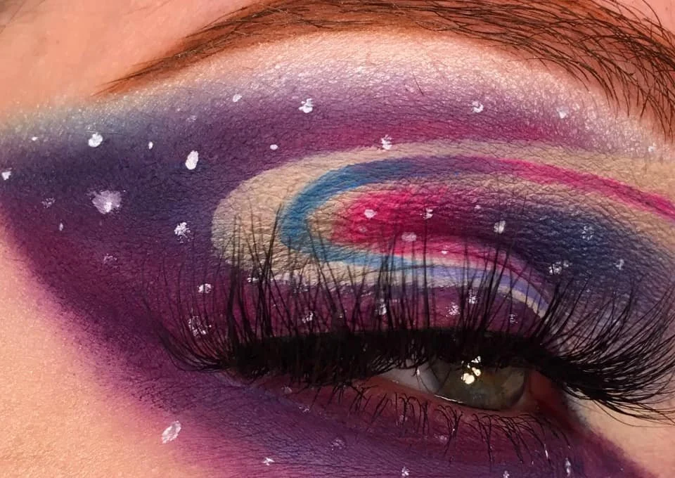 Close-up of eye with colorful galaxy-inspired eye makeup, including purple, pink, blue, and black shades, accented with white star-like dots and a sweeping pink line above the eyelid.