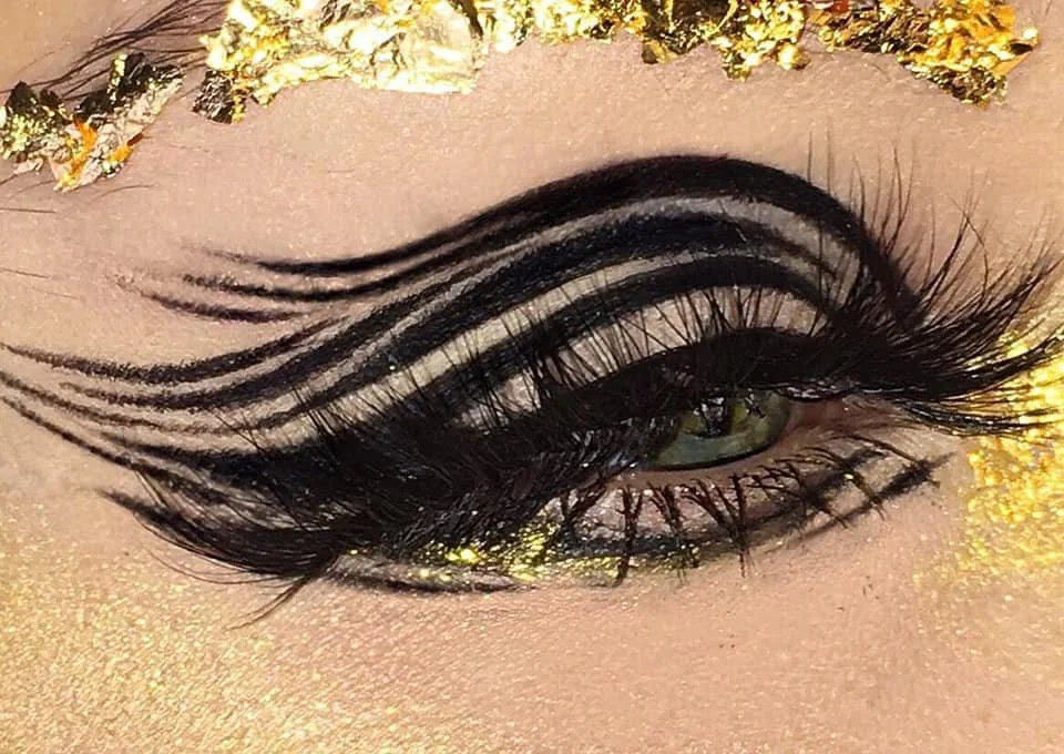 Close-up of a person's eye with elaborate black and white striped eyeliner, thick false eyelashes, and gold glitter makeup around the eye and on the eyebrow.