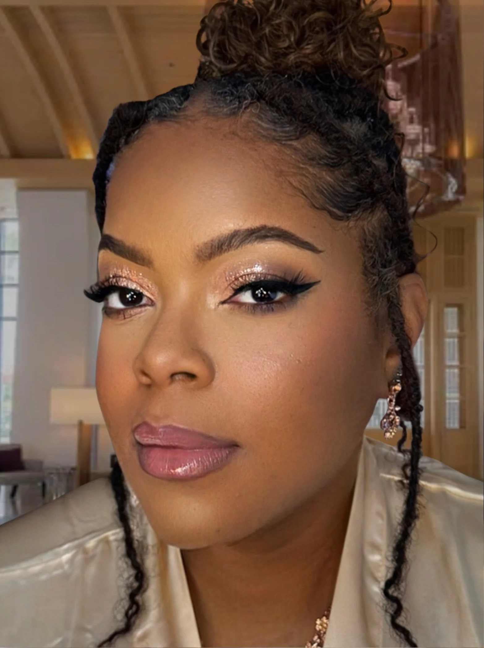 Close-up of a woman with styled hair, makeup including glittery eyeshadow, winged eyeliner, and glossy lips, wearing earrings and a light-colored top, indoors with a wooden ceiling.