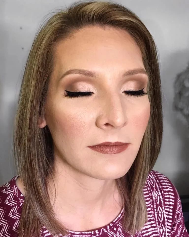 A woman with shoulder-length light brown hair, closed eyes, makeup including neutral eyeshadow, black eyeliner and mascara, and beige lipstick, wearing a maroon and white patterned top, standing against a light grey wall.