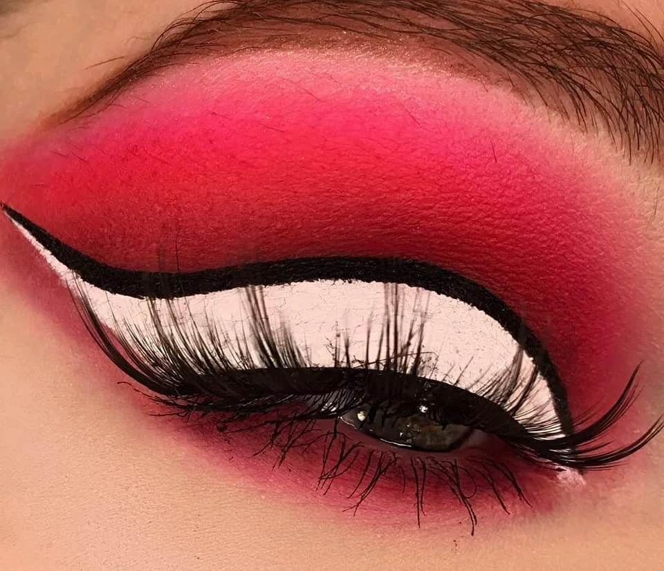 Close-up of an eye with vivid pink eyeshadow, bold black eyeliner, long dark eyelashes, and artistic white and black eye makeup design.