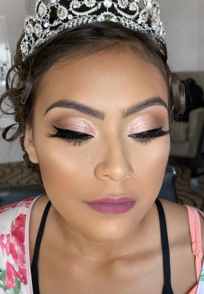 Close-up of a woman with a glittery pink eyeshadow, black winged eyeliner, long lashes, and pink lipstick. She wears a silver tiara with intricate designs and gemstones, and a floral-patterned garment.