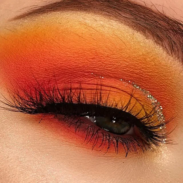 Close-up of a person's eye with vibrant orange and yellow eyeshadow, black eyeliner, and rhinestones along the eyelid.