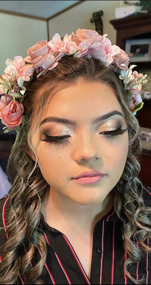 Young woman with closed eyes wearing a floral crown of pink and cream roses, and makeup with shimmering eyeshadow and long lashes.