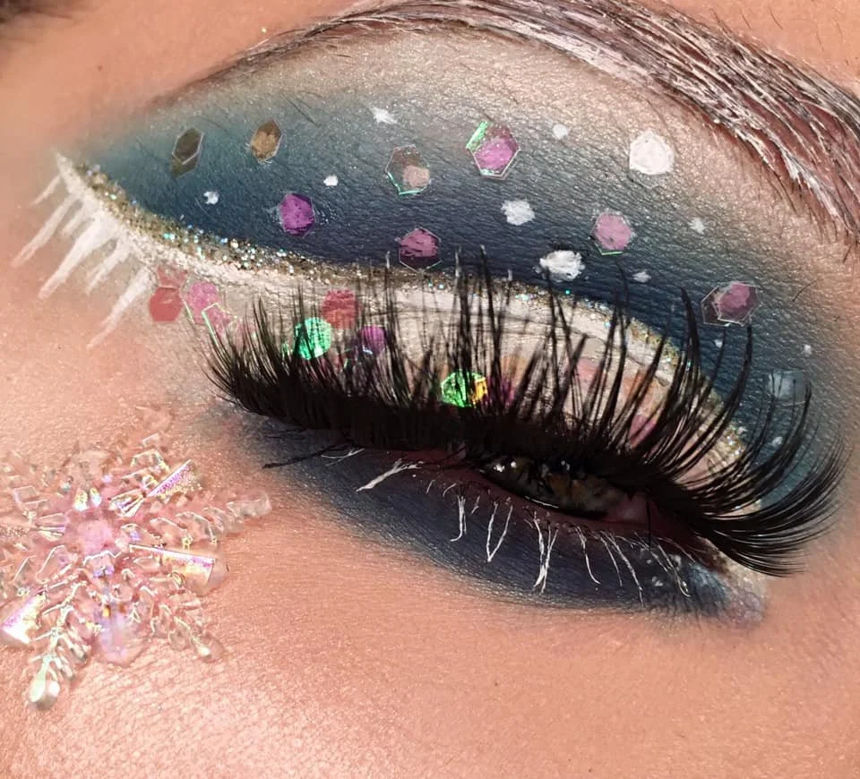 Close-up of an eye with elaborate makeup, including blue eyeshadow, white and pink accents, and shiny sequins. Long eyelashes and a decorative snowflake sticker are also visible.