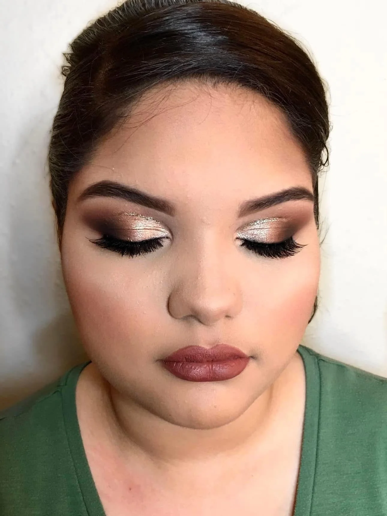 Close-up of a woman with dark hair and makeup, wearing a green top, with her eyes closed, showing eyeshadow and long eyelashes.