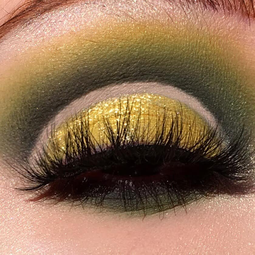 Close-up of a closed eye with yellow glitter eyeshadow, dark black mascara, and eyeshadow in green, black, and yellow shades.