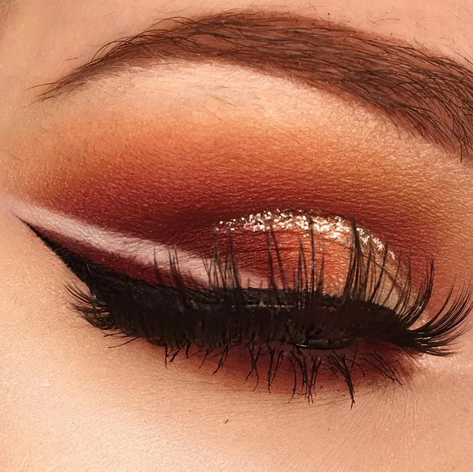 Close-up of a person's closed eye with warm-toned eyeshadow, black eyeliner, false eyelashes, and glitter on the eyelid.