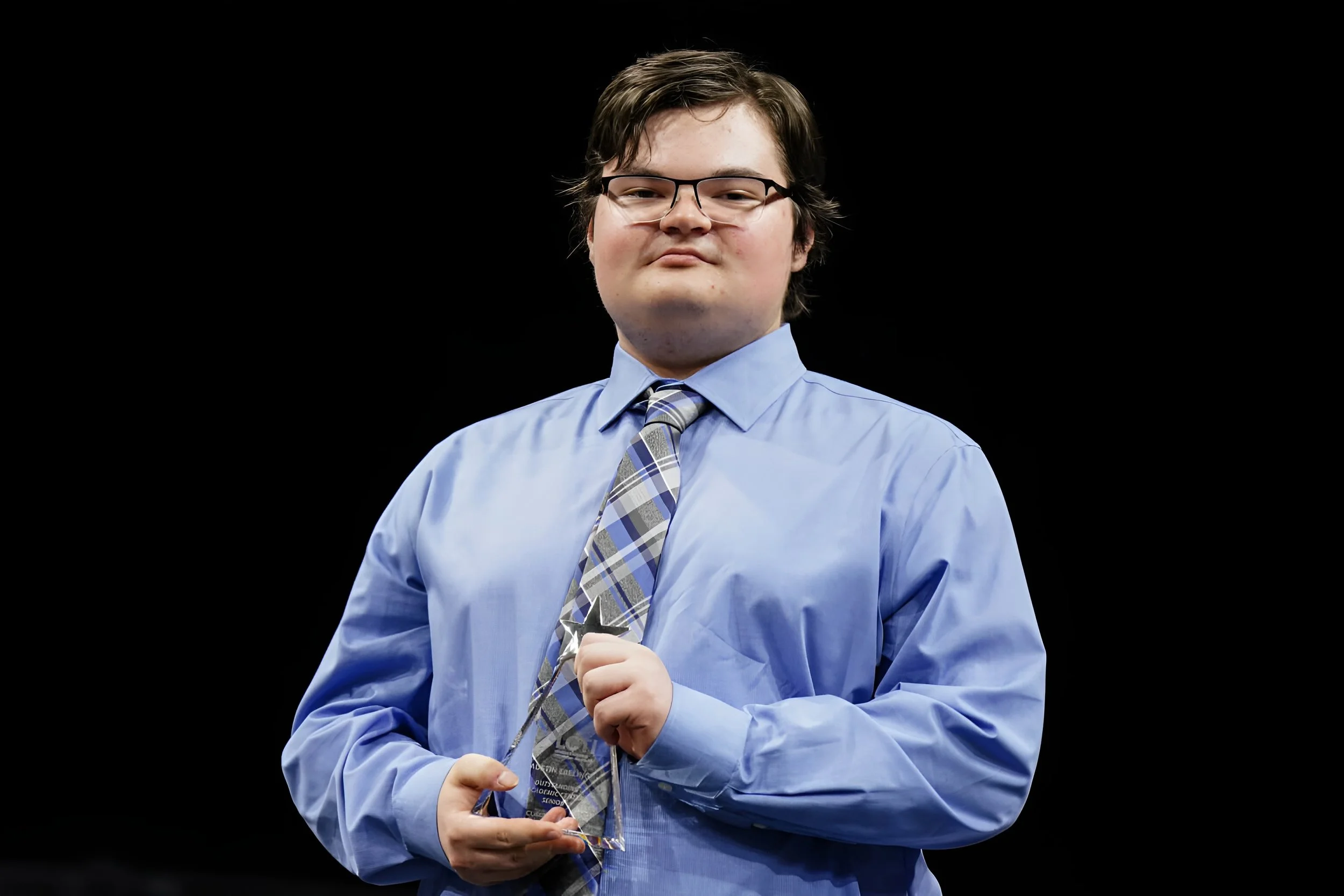 Austin Ebeling in a blue dress shirt and plaid tie holding a glass award, standing against a black background.