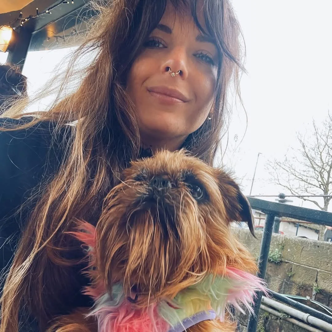 A woman with long, wavy brown hair and a septum piercing holding a small, wet dog with colorful, fluffy feathers around its neck outdoors.
