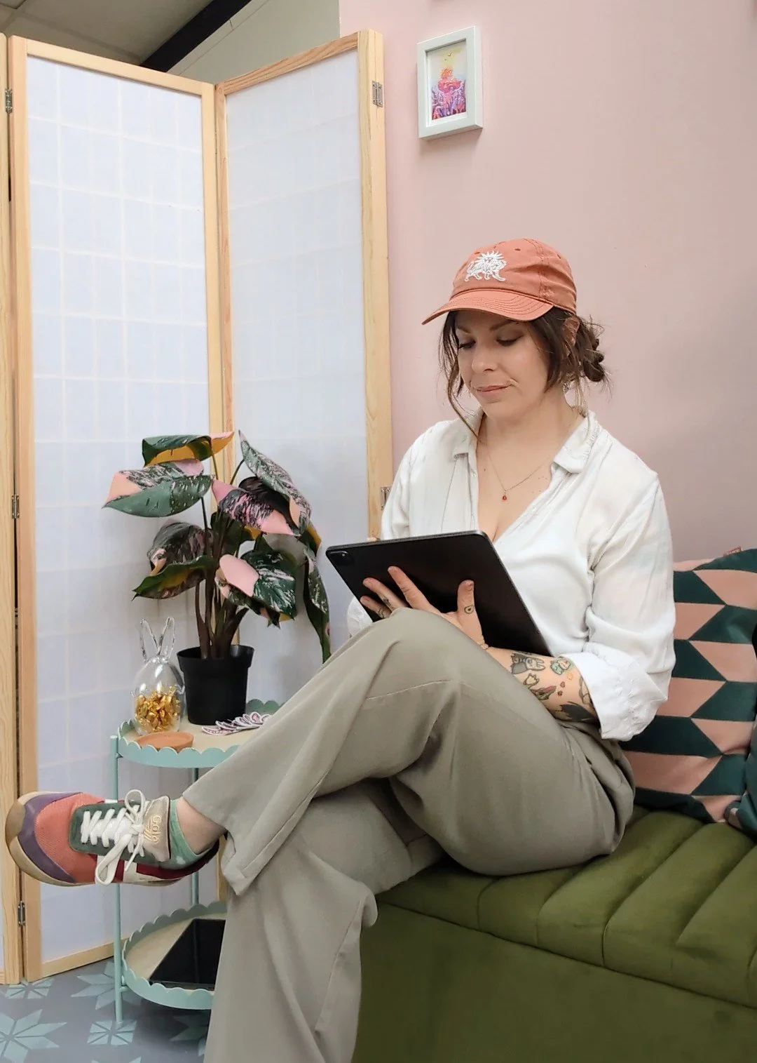 A woman sitting on a green sofa, wearing a beige baseball cap, white blouse, beige pants, and colorful sneakers, holding a tablet in her hands. The room has pink walls, a folding wooden screen, a potted plant, and a small table with decorative items including a glass bunny and a plate of chocolates.