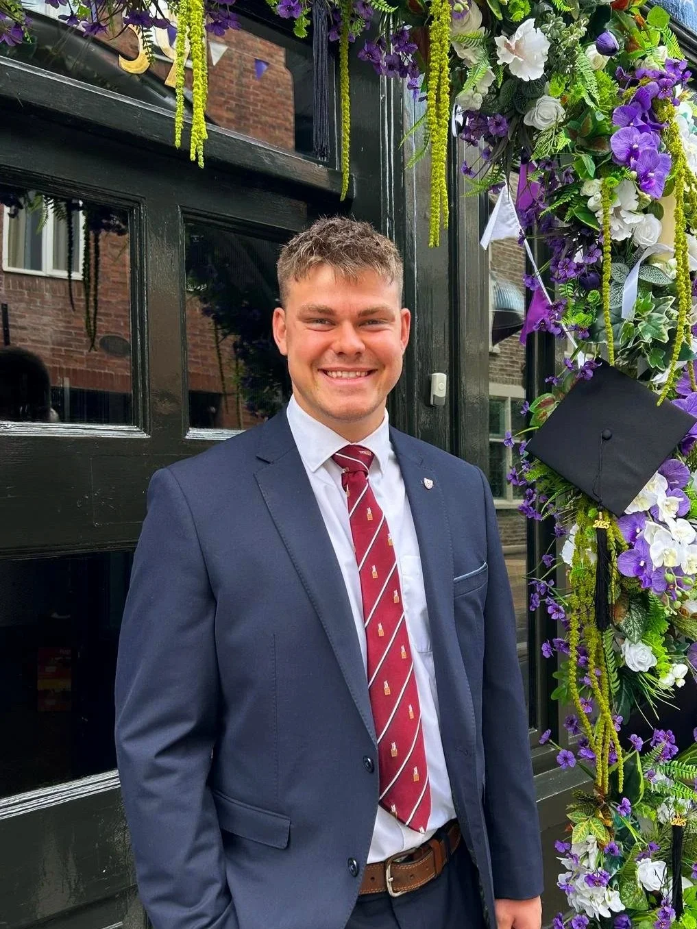 Young man in a navy suit with a red tie, smiling at a graduation ceremony, standing beside decorated floral arch with purple and white flowers and a black graduation cap hanging on a ribbon.