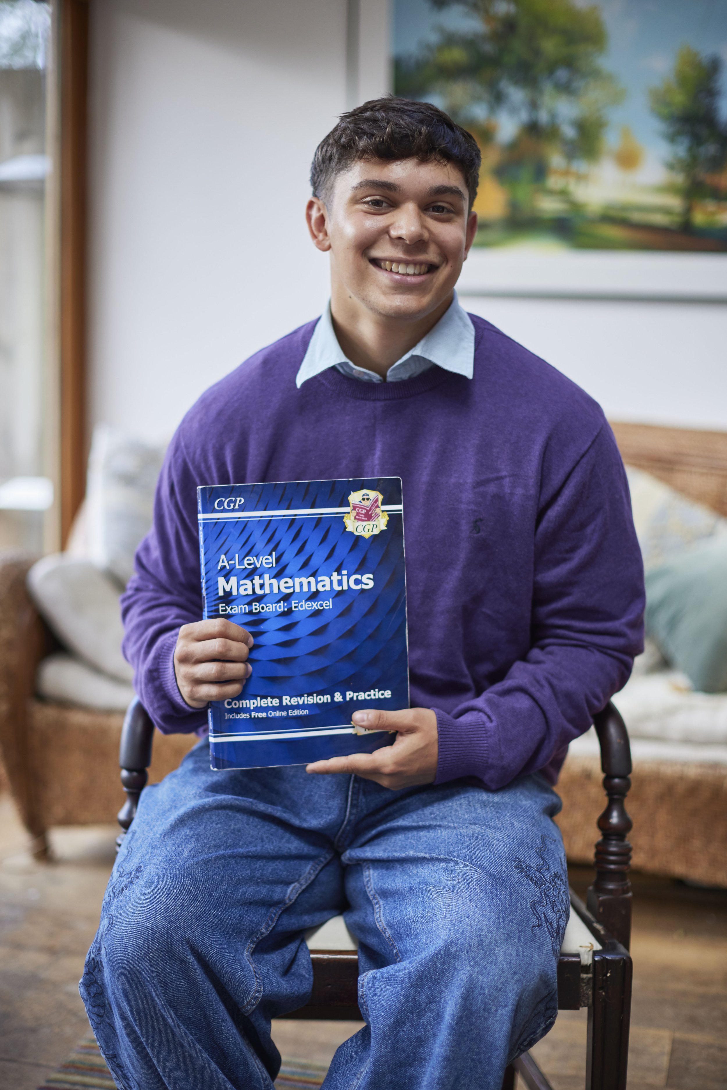 A young man smiling while sitting on a chair, holding a mathematics study guide book labeled 'A-Level Mathematics Complete Revision & Practice'.