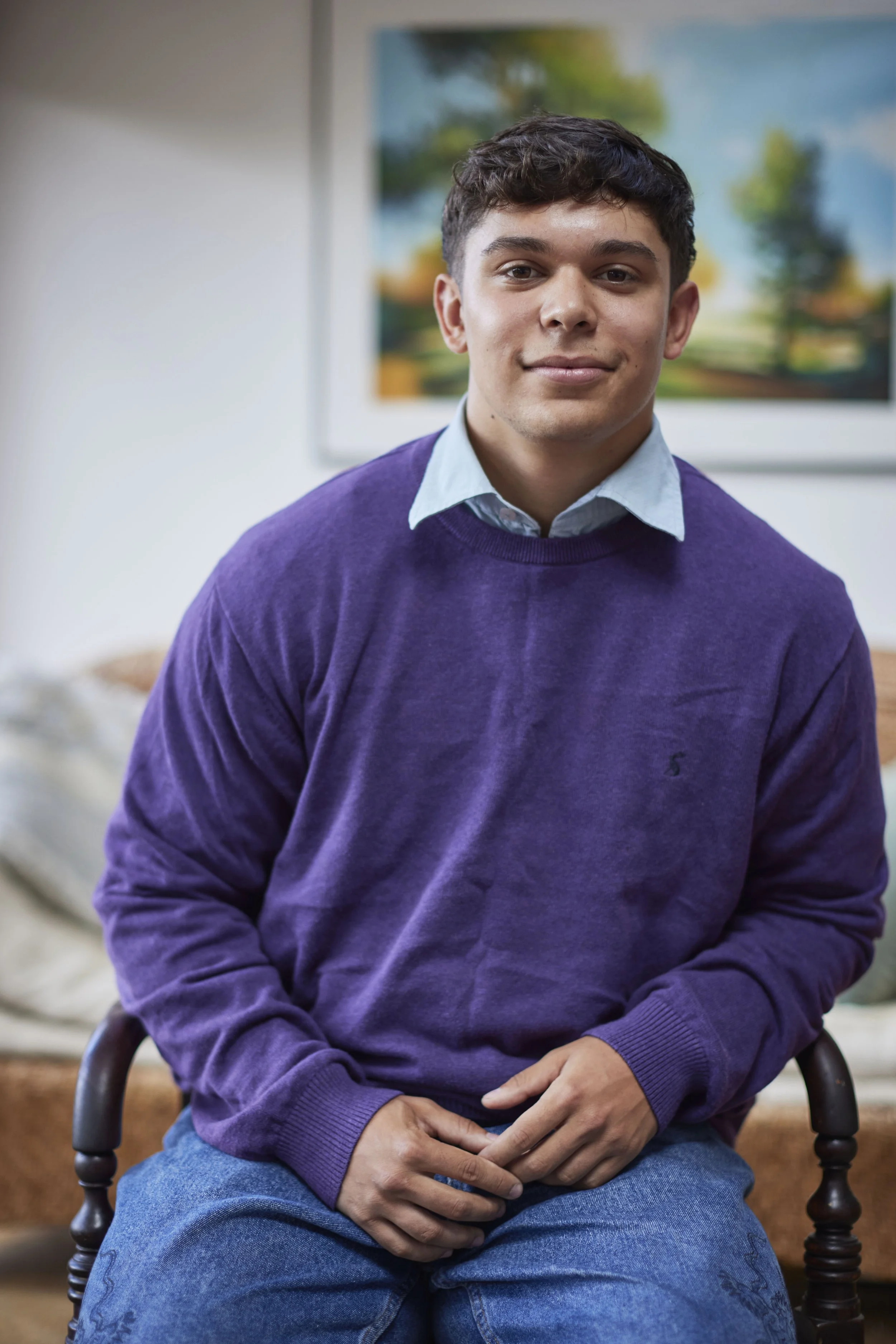 A young man sitting on a wooden chair indoors, wearing a purple sweater and a light blue collared shirt. There is a blurred landscape painting on the wall behind him.