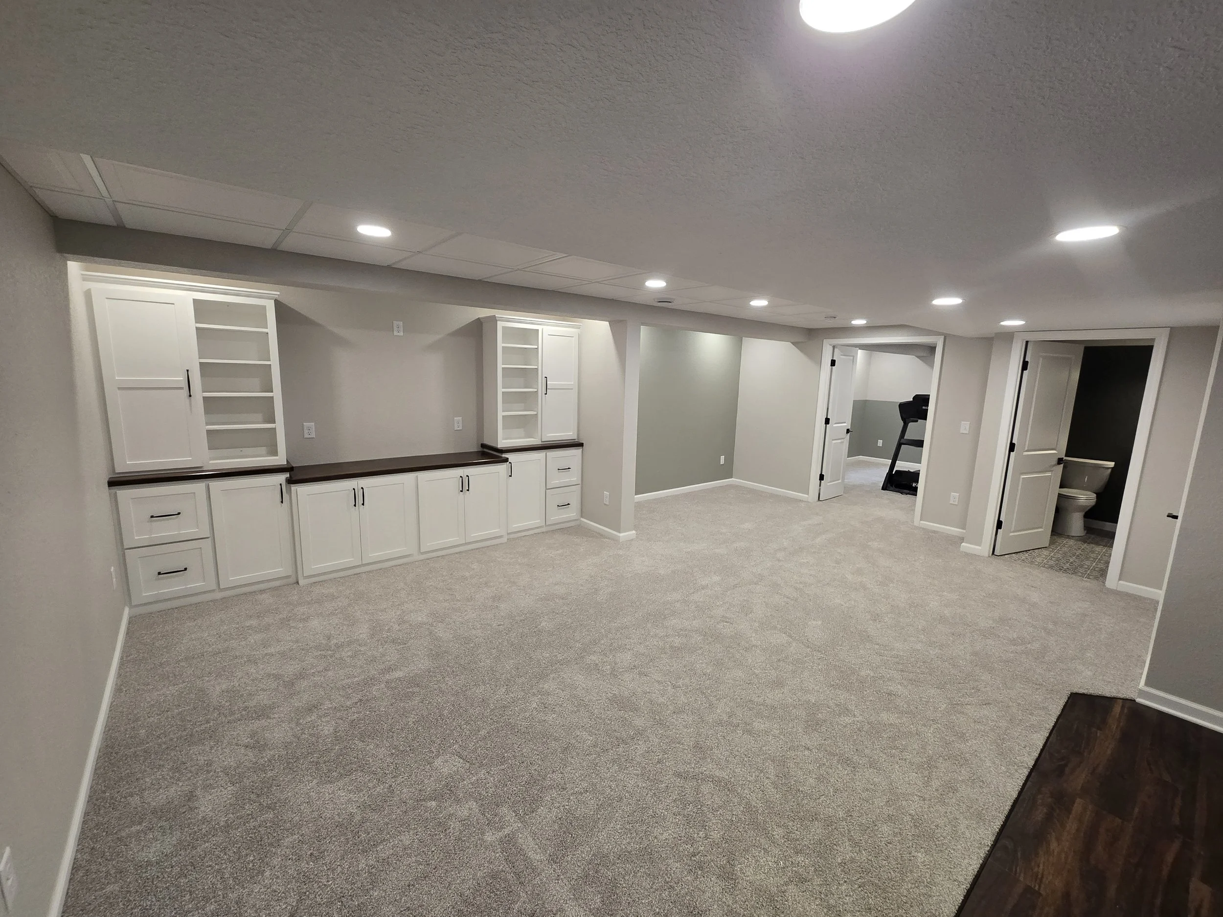 Empty finished basement with beige carpet, built-in white cabinets and shelves, multiple ceiling lights, and three open doorways leading to small rooms including a bathroom and a workout area.