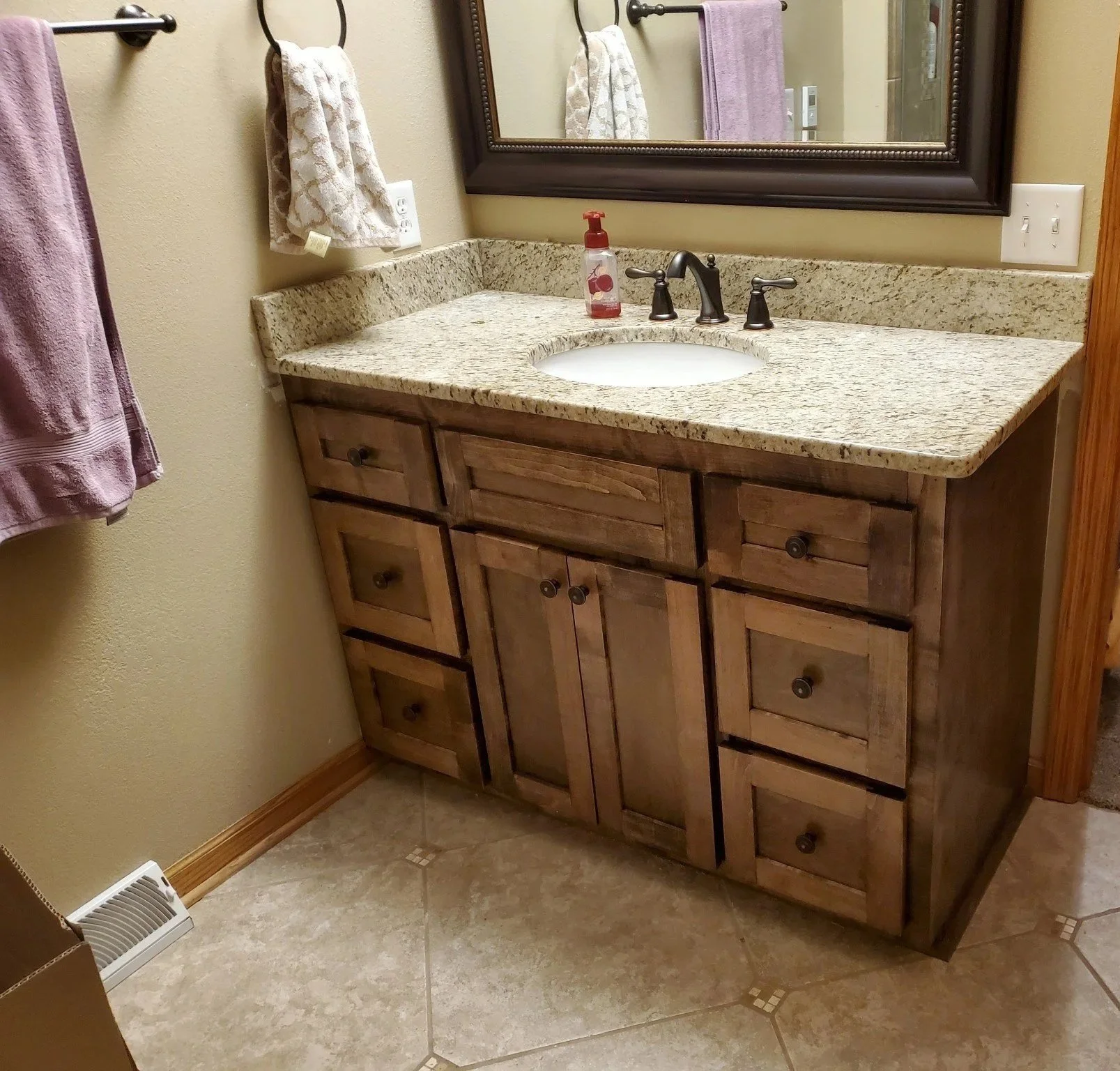 Bathroom vanity with a granite countertop, a mirror, a soap dispenser, and a wooden cabinet with multiple drawers. Part of a bedroom and a person standing nearby are visible in the background.