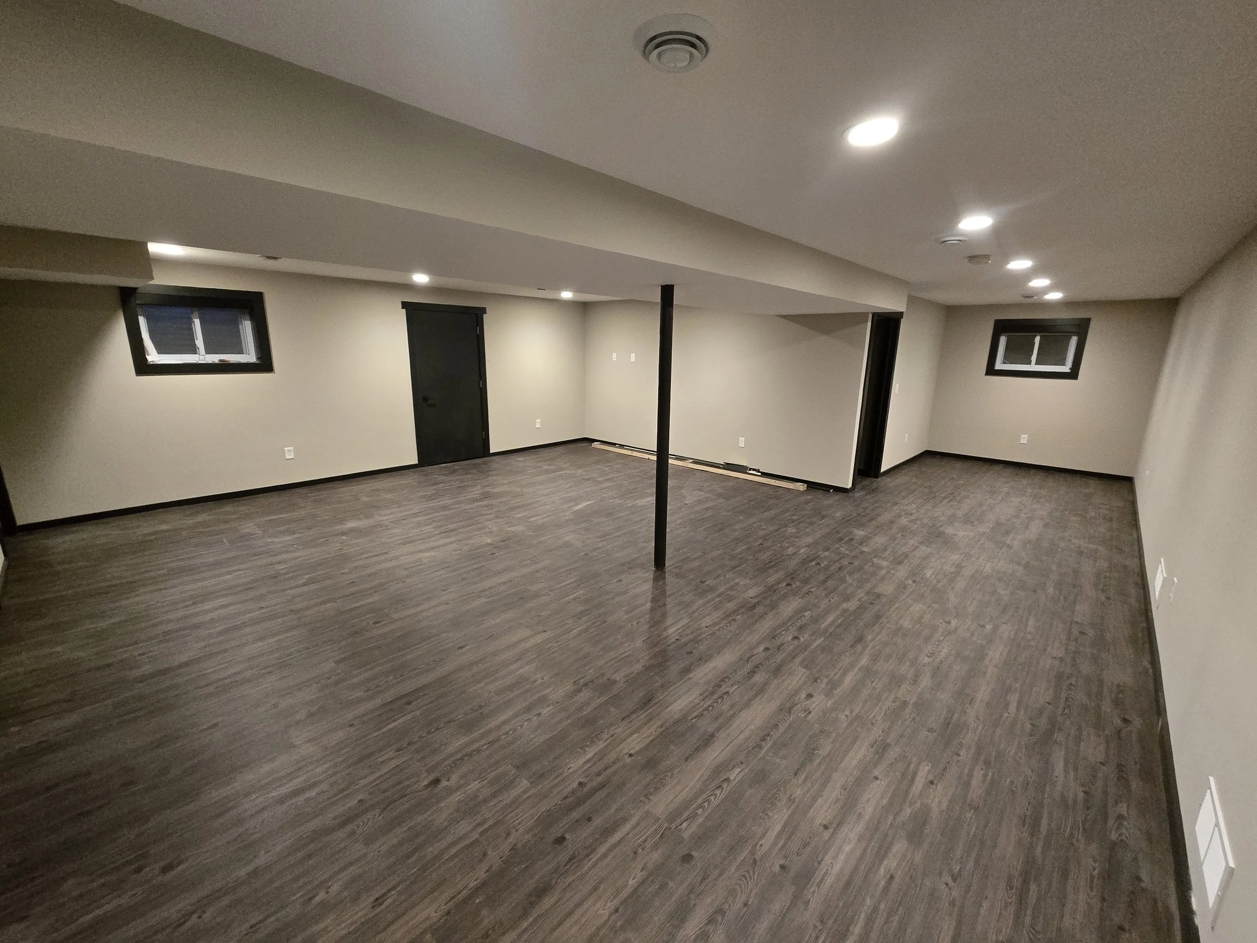 An empty basement room with gray walls, dark wood flooring, small windows, ceiling lights, and a black door.