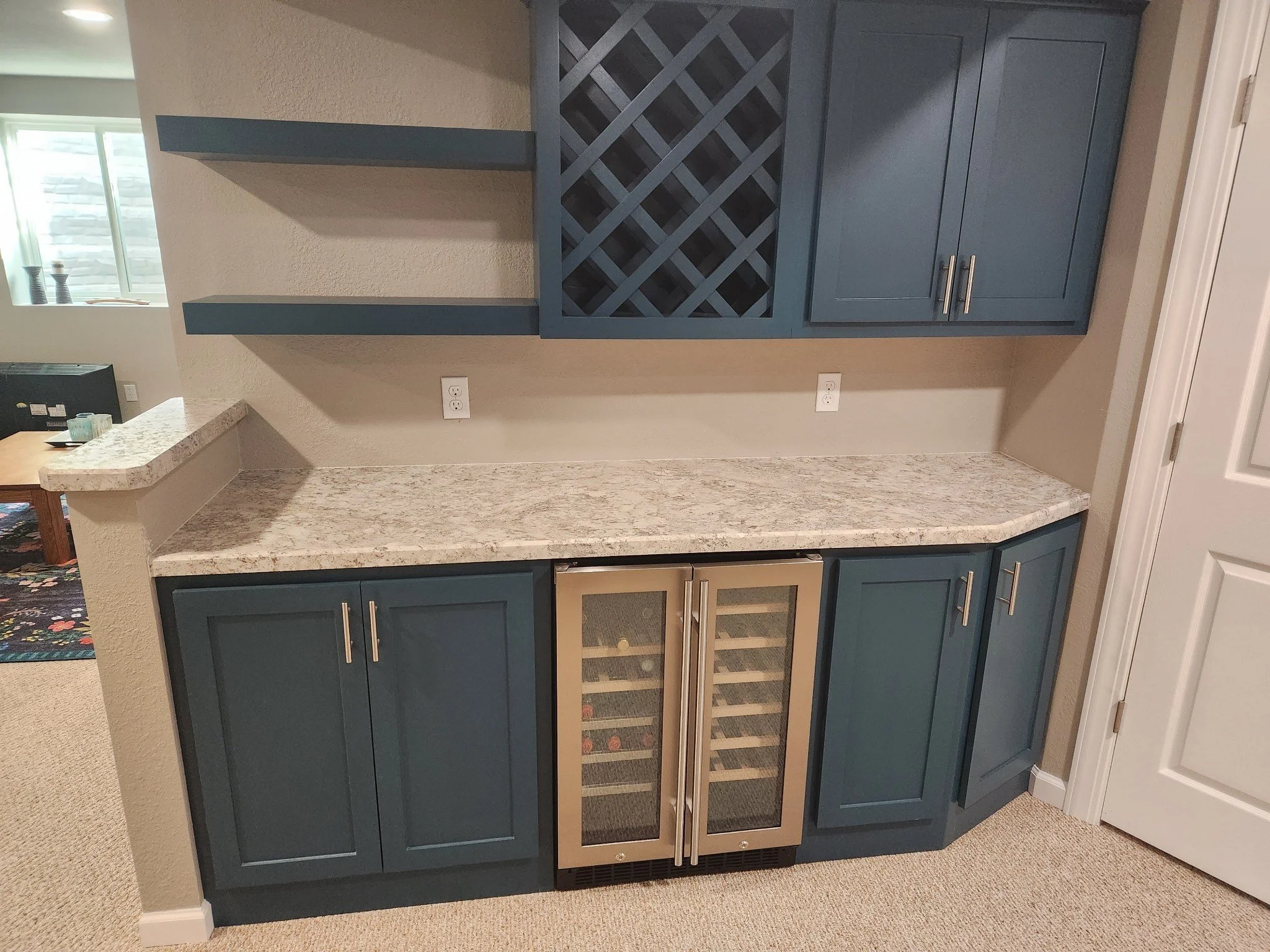 A home bar area with blue cabinets, a granite countertop, and a built-in wine cooler, with open space on the left and electrical outlets on the wall.