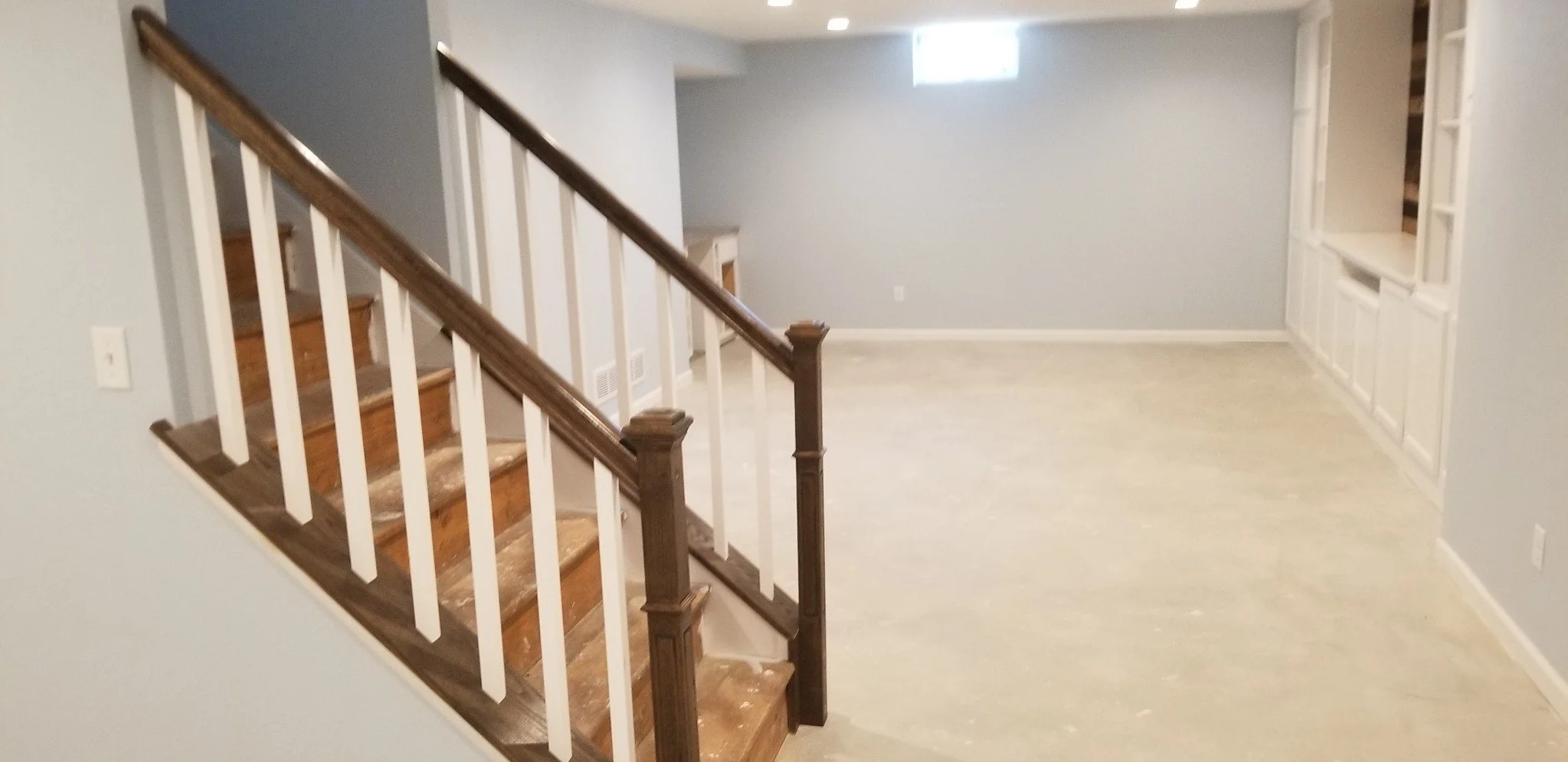Empty living room with light gray walls, wood staircase with white balusters, built-in white shelves, and a small window near the ceiling.