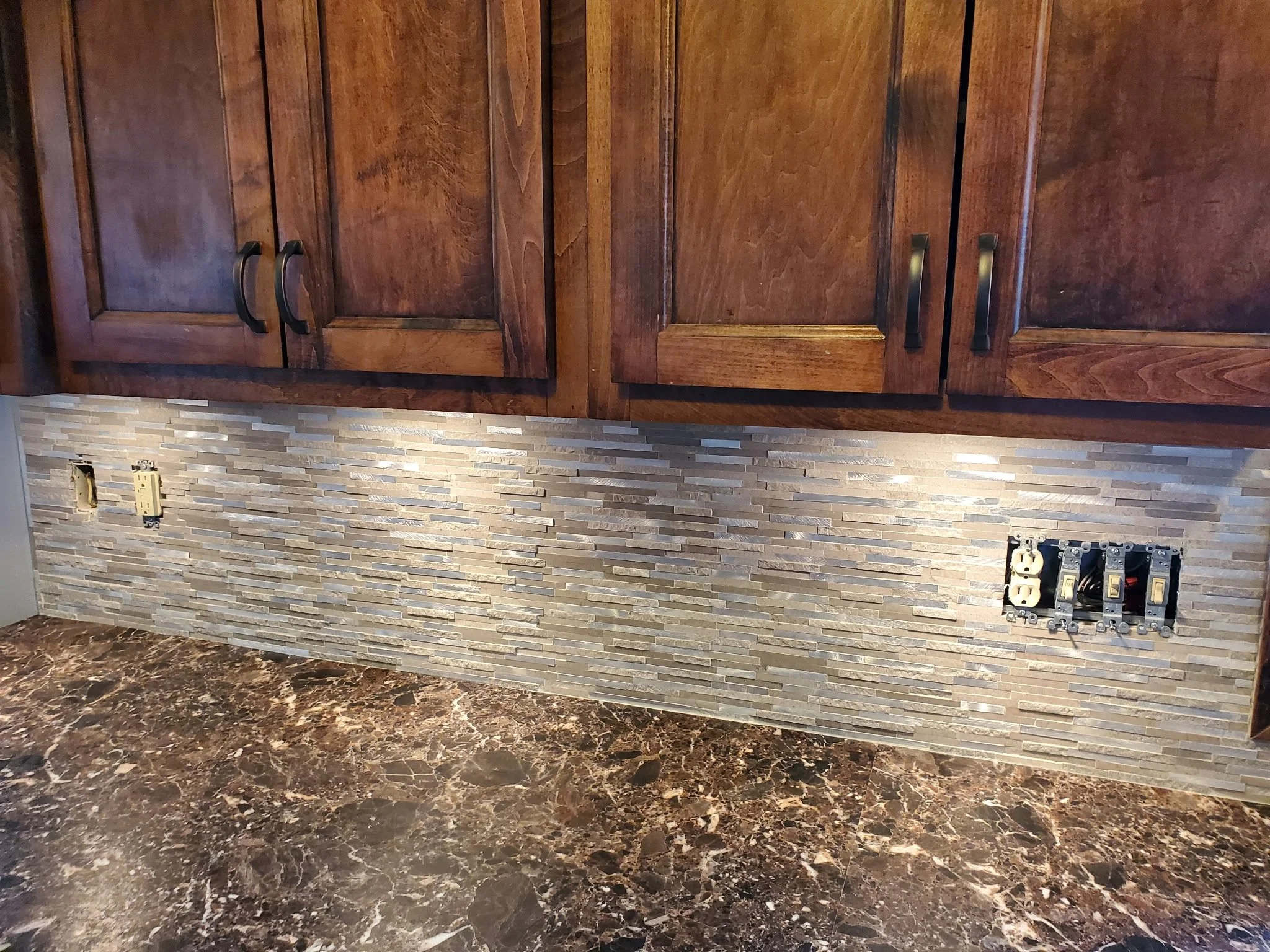 Kitchen countertop with empty electrical outlets and switches, wooden cabinets above, and a textured tile backsplash.