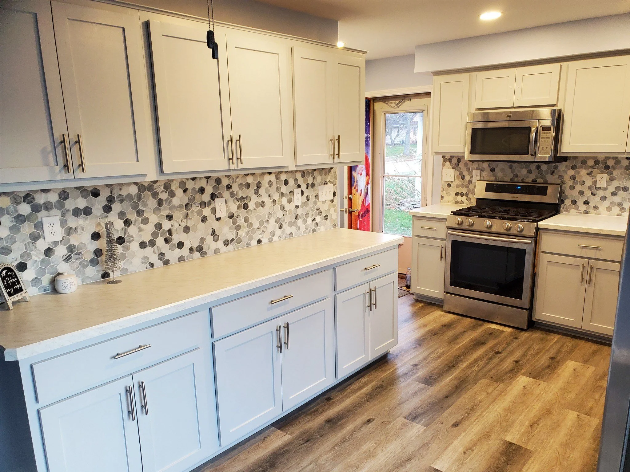 Kitchen with white cabinets, a hexagonal tile backsplash, stainless steel oven and microwave, wooden flooring, and a door leading outside.