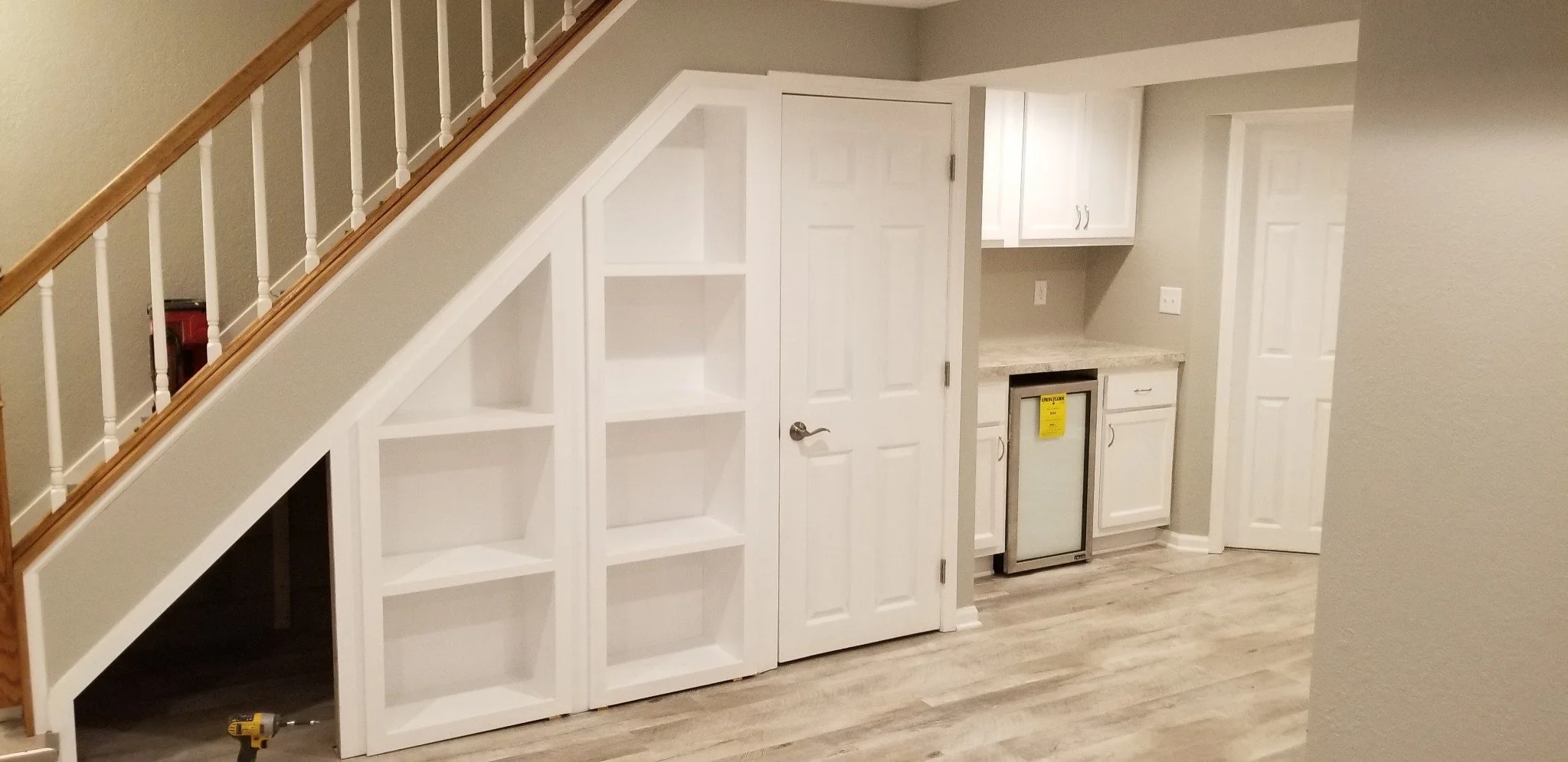Interior of a room with a staircase on the left, built-in white shelves underneath, a white door in the center, and a small kitchenette with white cabinets, granite countertop, and mini fridge with a yellow sticker on it on the right.