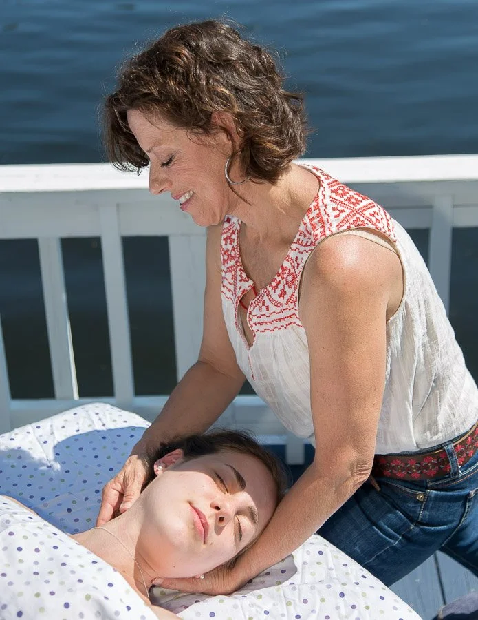 A woman giving a massage to another woman lying on a bed outdoors near water.