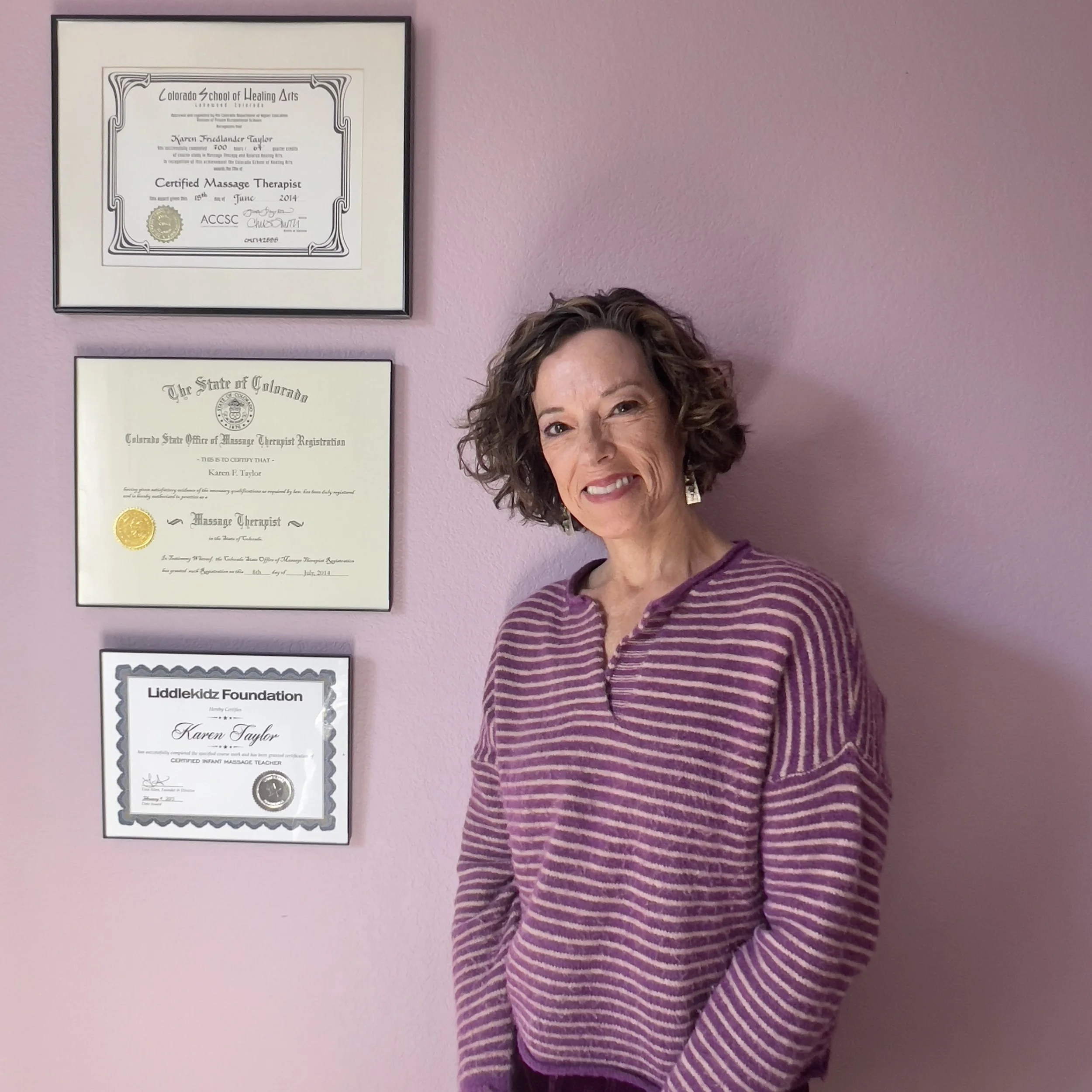 A woman with curly hair, wearing a purple striped sweater, standing in front of a pink wall with three framed certificates hanging on it.
