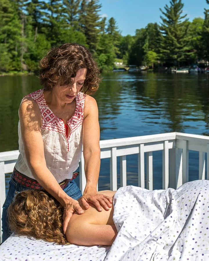 A woman giving a massage to a person lying on a bed outdoors by a lake with green trees in the background.