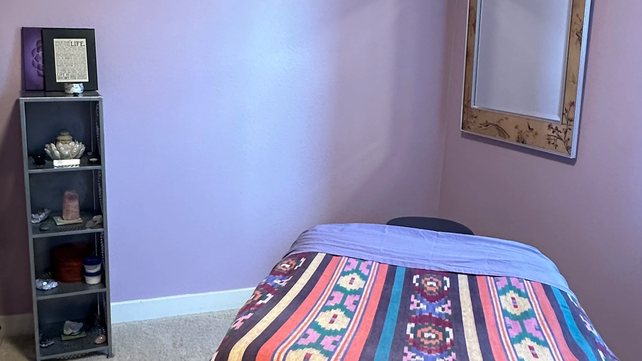 A bedroom with a bed covered with a colorful patterned quilt, a small gray shelf with decorative items, and a framed mirror on a pink wall.