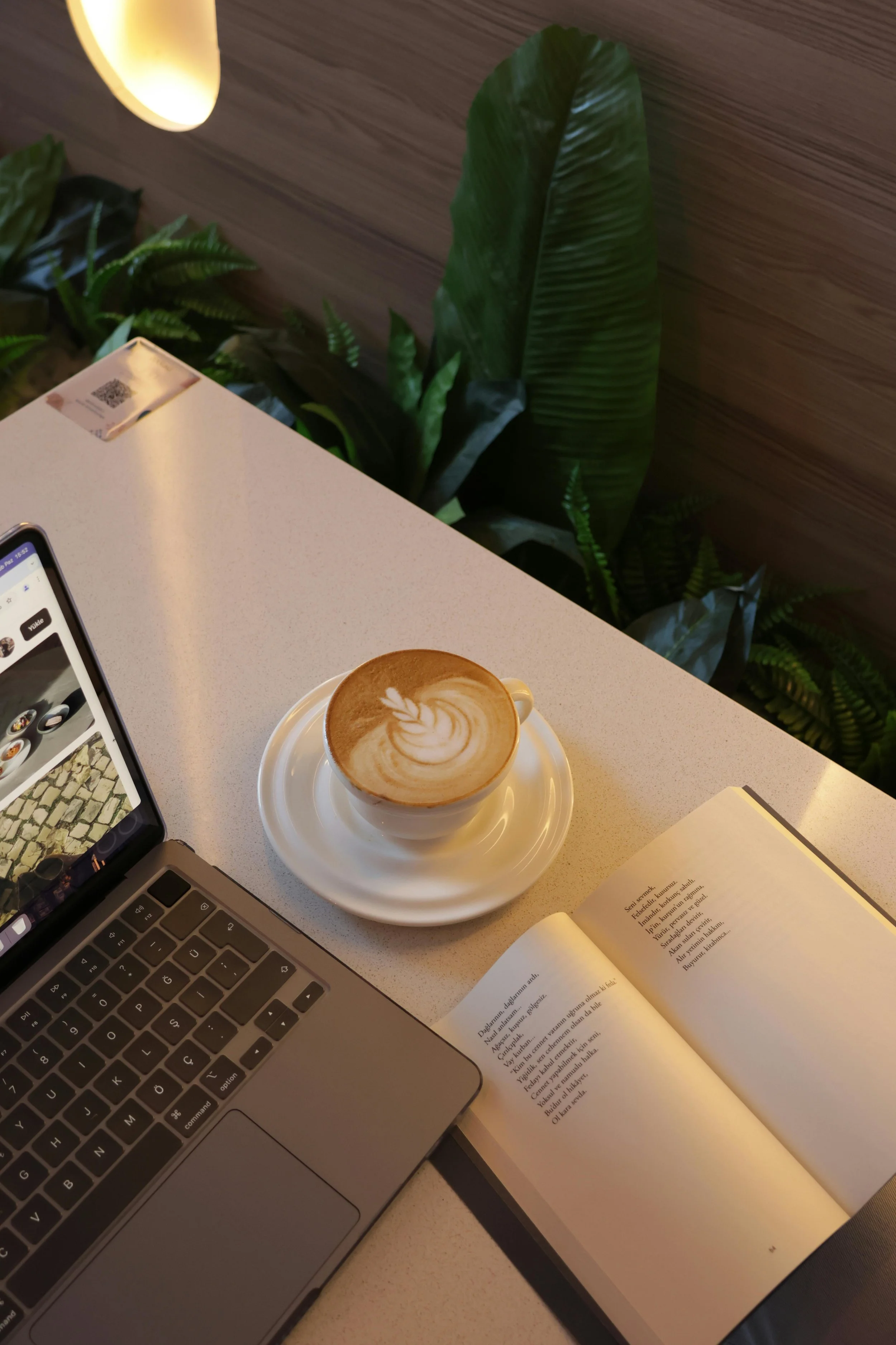 A cup of latte with latte art on a white saucer, partially open laptop, open book, and a QR code on the table in a cafe with green plants and wooden wall decor.