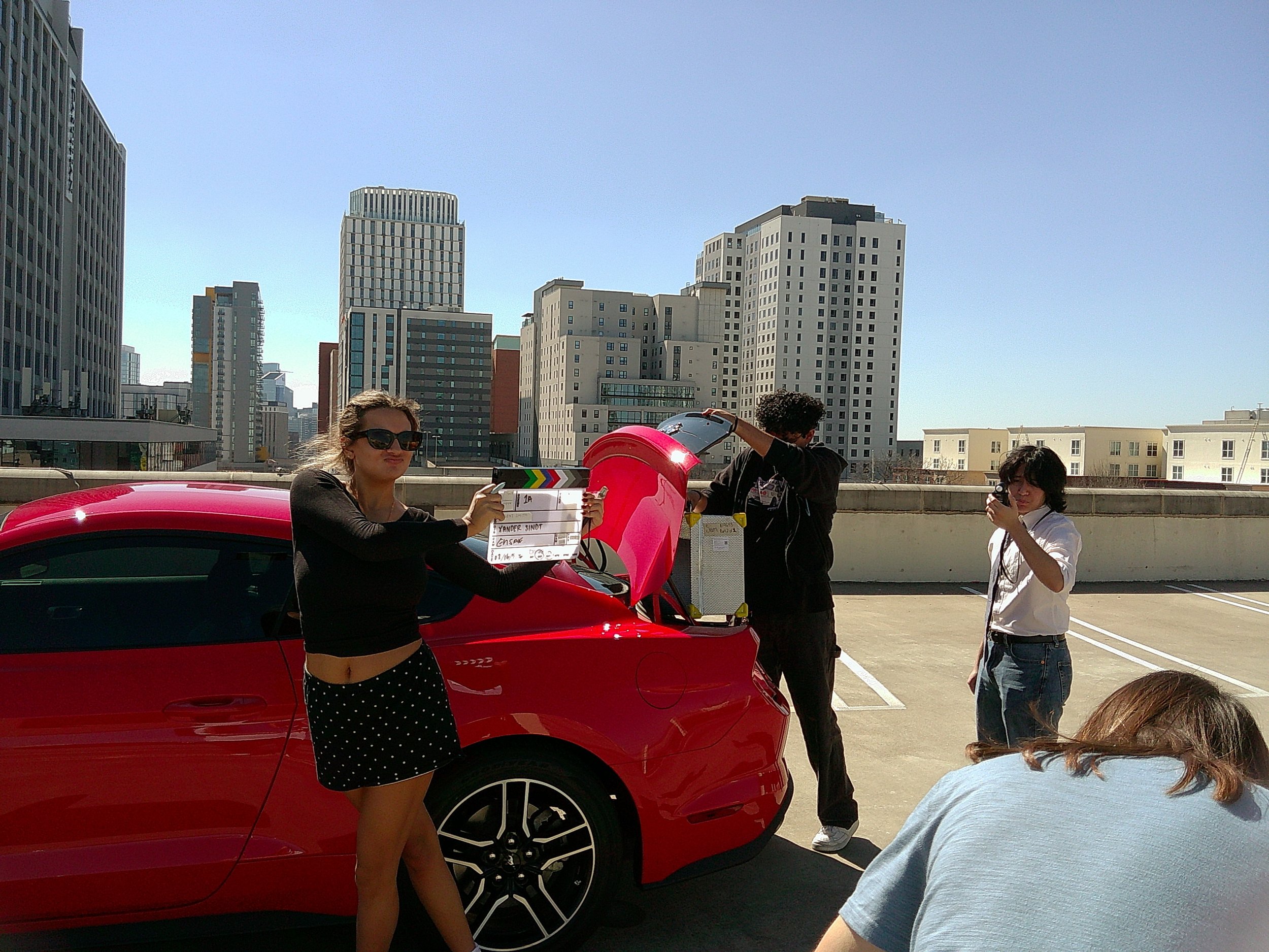 People standing around a red sports car on a rooftop with a city skyline in the background, one person holding a film slate.
