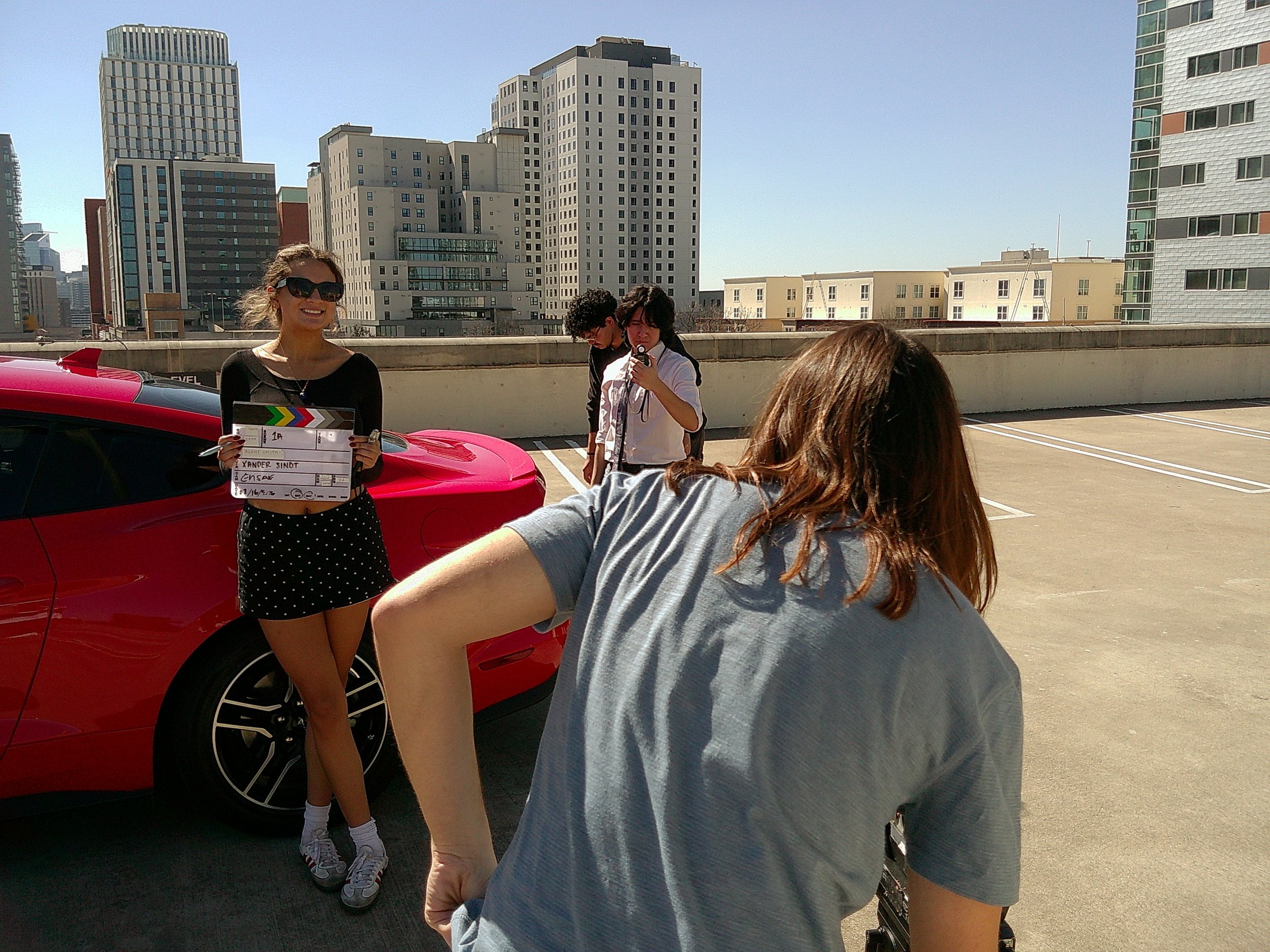 A woman holding a clapperboard standing next to a red sports car on a rooftop parking lot with city buildings in the background. There are a few other people around, one taking a photo and others talking.