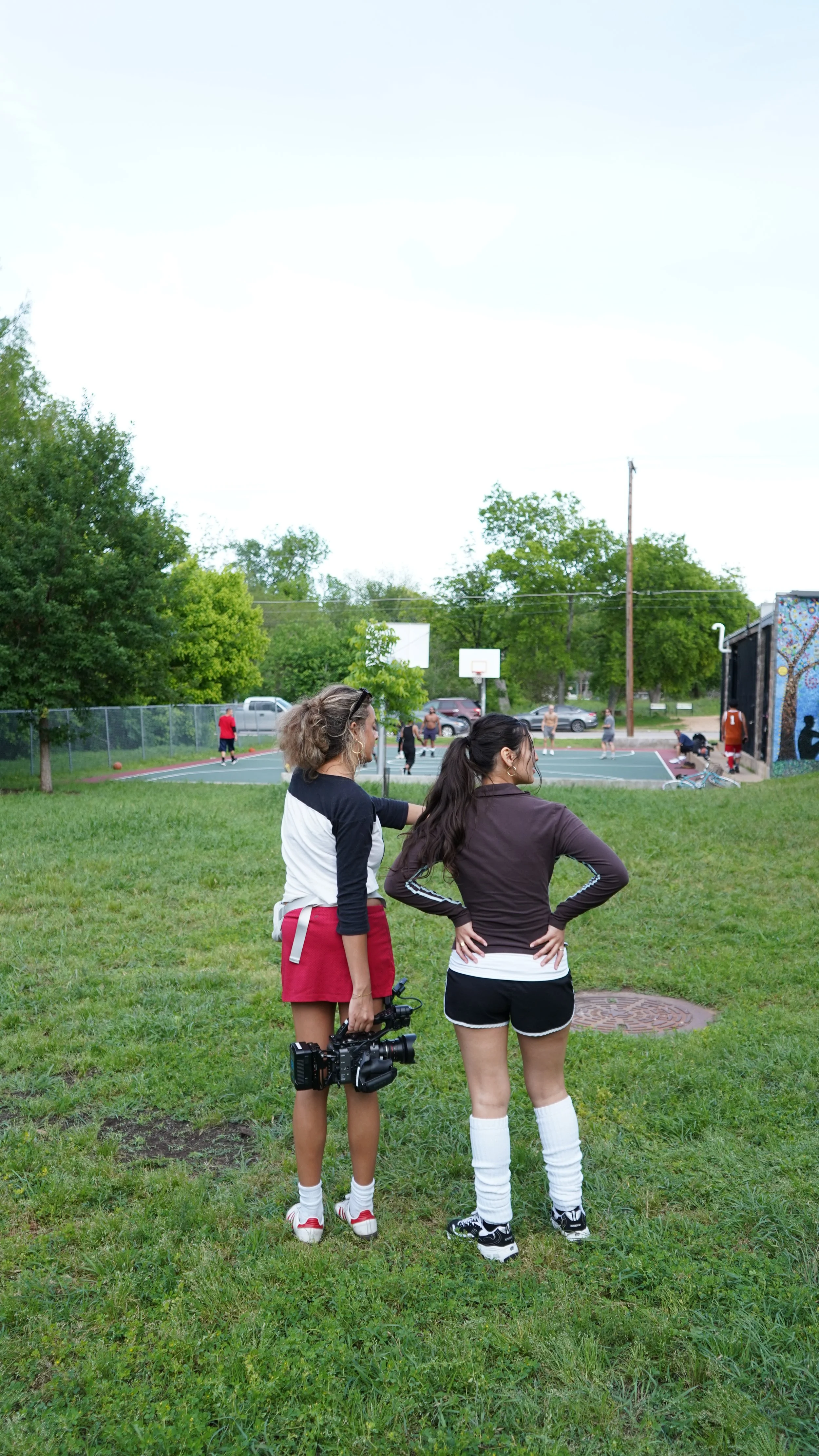 Two women stand on a grassy area watching a basketball game at an outdoor court, surrounded by trees and a chain-link fence, with people playing basketball in the background.
