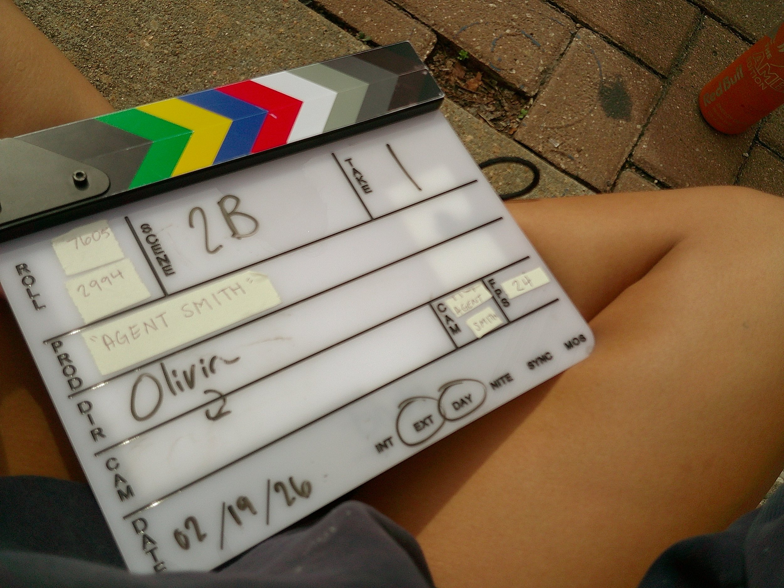 Close-up of a clapperboard with handwritten notes, a colorful sticker on the top part, and a weathered brick surface in the background. A person's arm is partially visible in the foreground, and a red Red Bull can is on the ground to the right.
