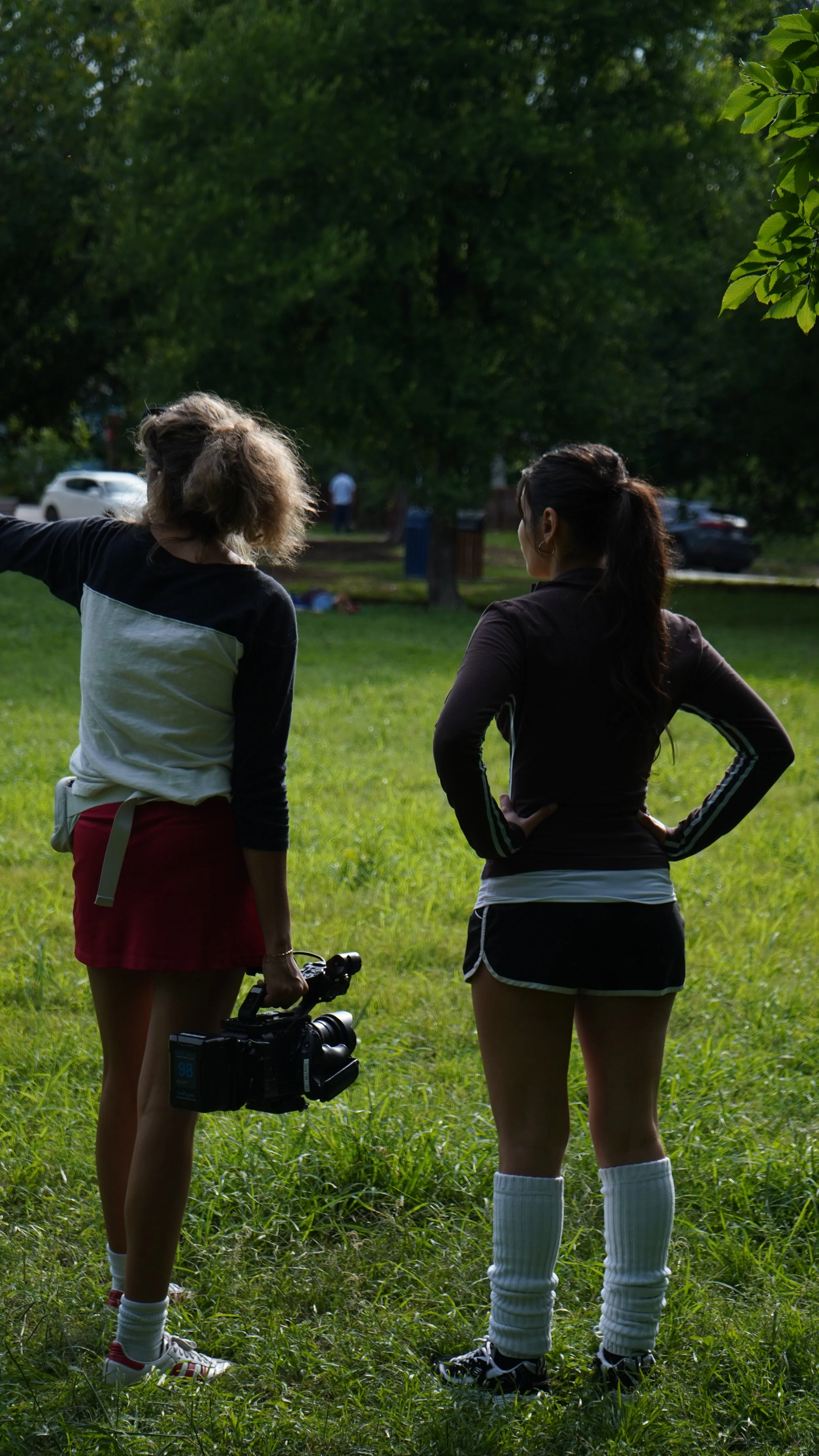 Two women standing in a grassy park, one holding a camera, the other with hands on hips, looking at something in the distance, with trees and parked cars in the background.