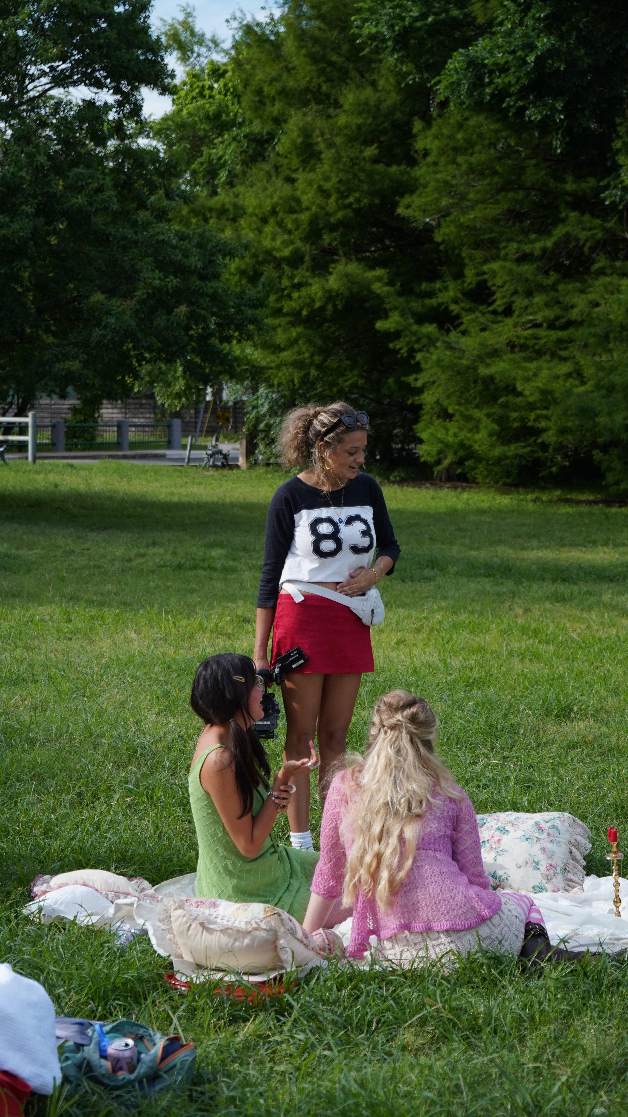 A woman in a baseball-style shirt with the number 83, standing on a grassy field, surrounded by children sitting on blankets, outdoors with trees in the background.
