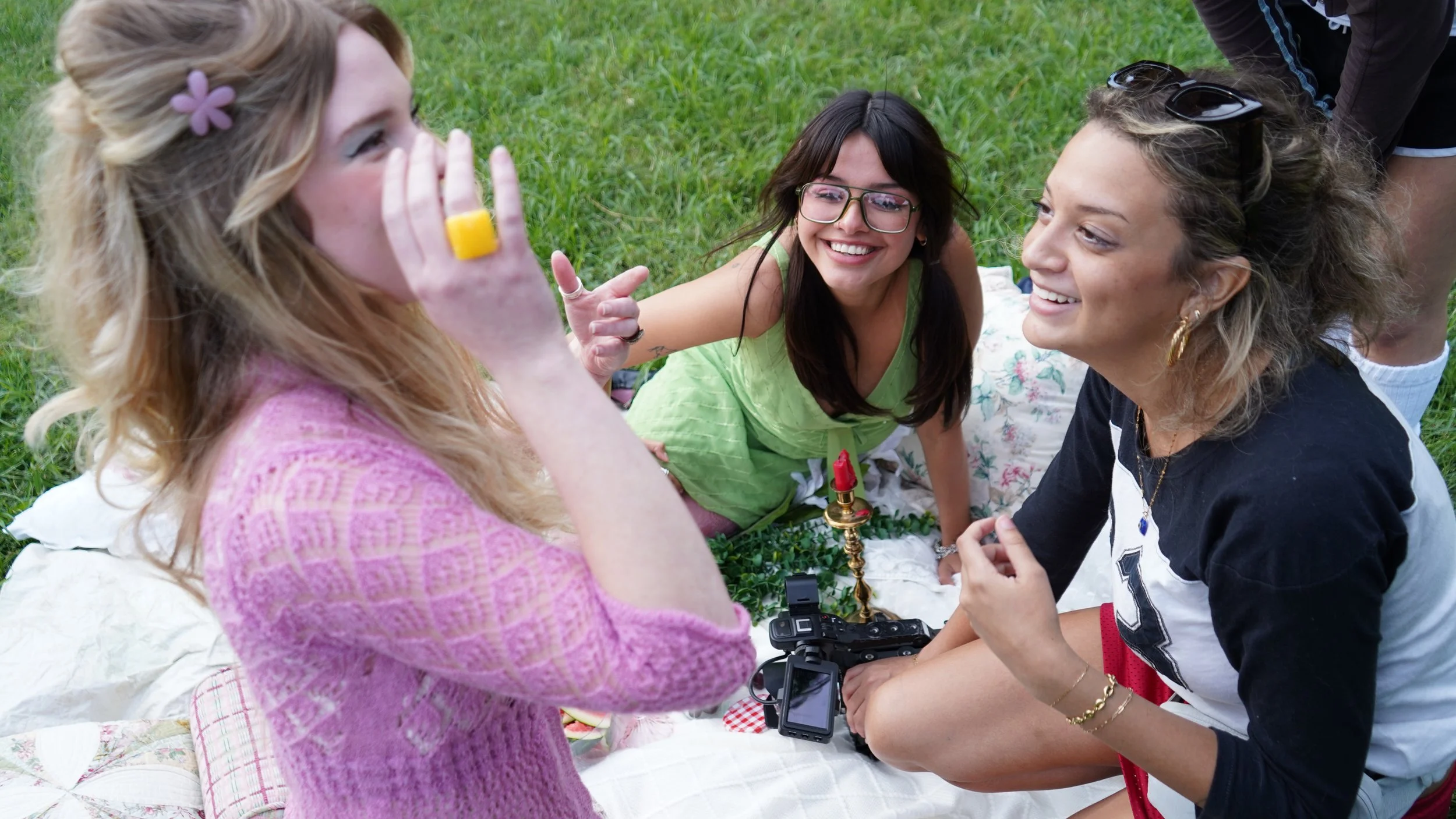 Four women sitting on a blanket on the grass, smiling and engaging in a conversation, with one woman holding a camera and another pointing.
