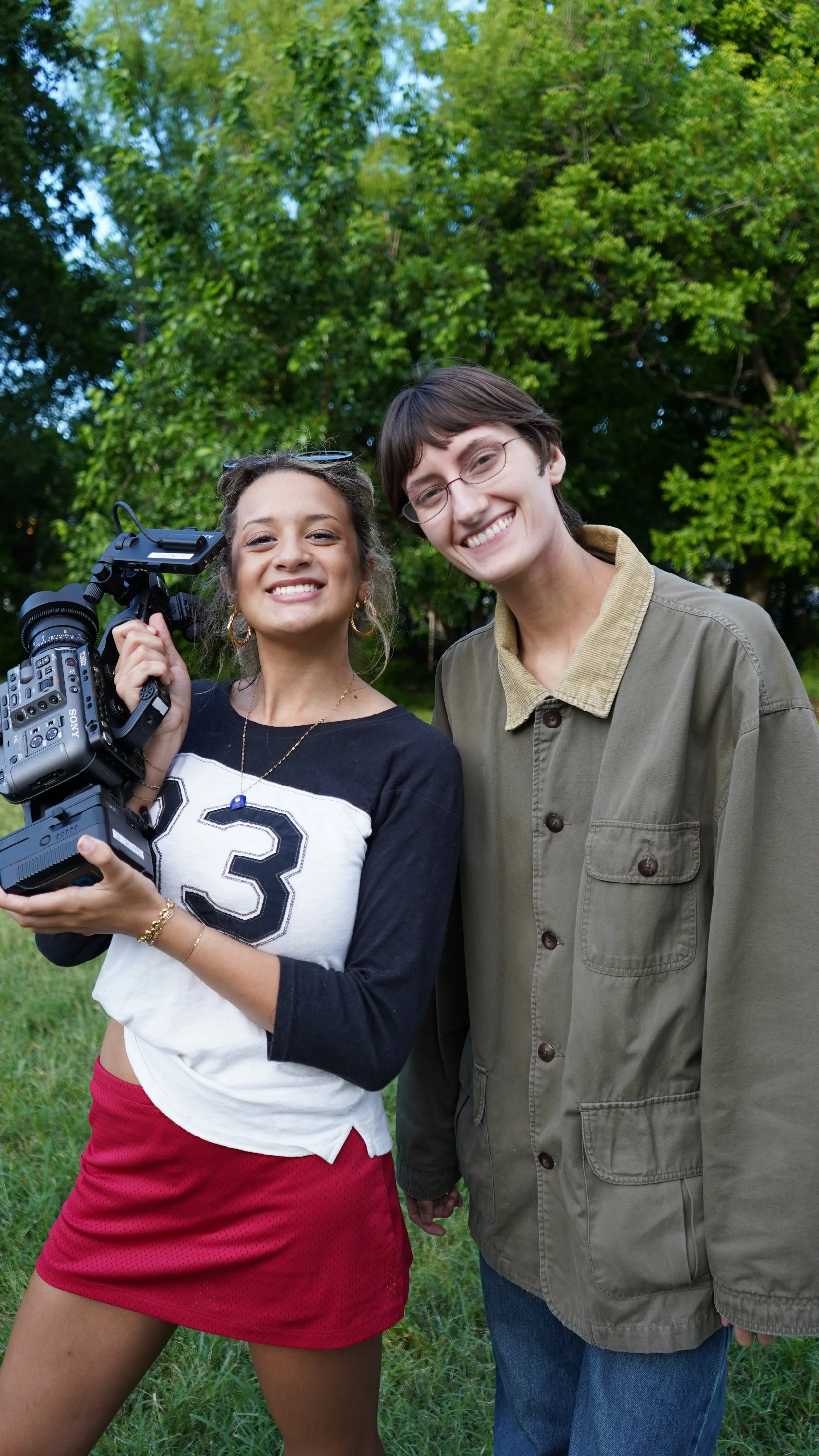 A young woman holding a professional video camera and a young man with glasses smiling outdoors in front of green trees.