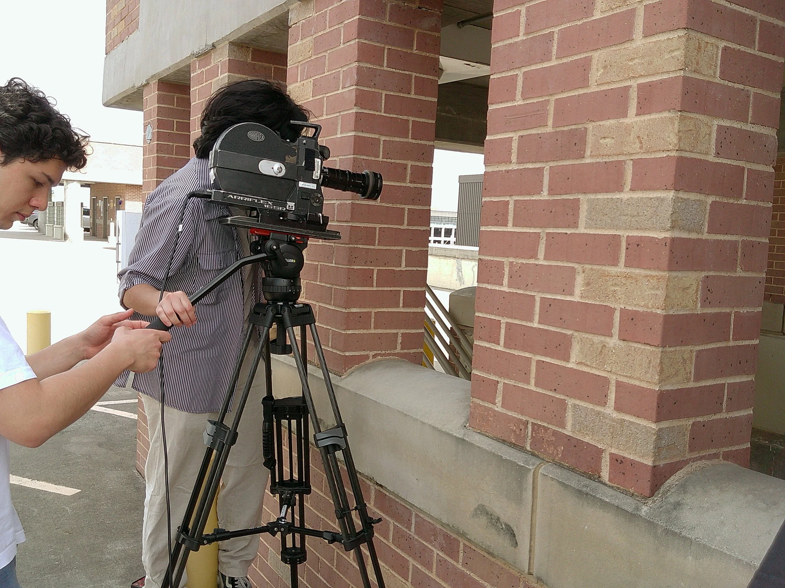 Two people working with surveying equipment on a brick building exterior. The person in focus is operating a theodolite on a tripod, while another person is assisting or taking notes.